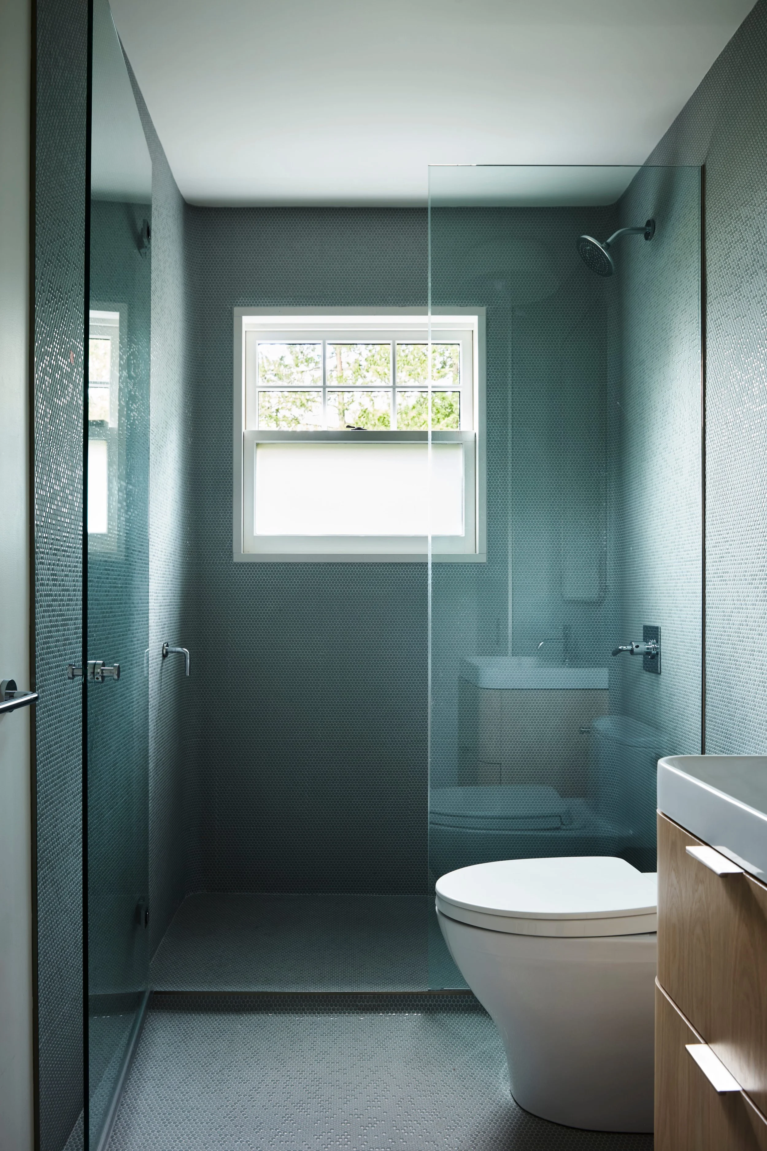 Modern bathroom with gray textured walls, a window, a walk-in shower with a glass door, a wall-mounted showerhead, and a white toilet with a wooden vanity nearby.