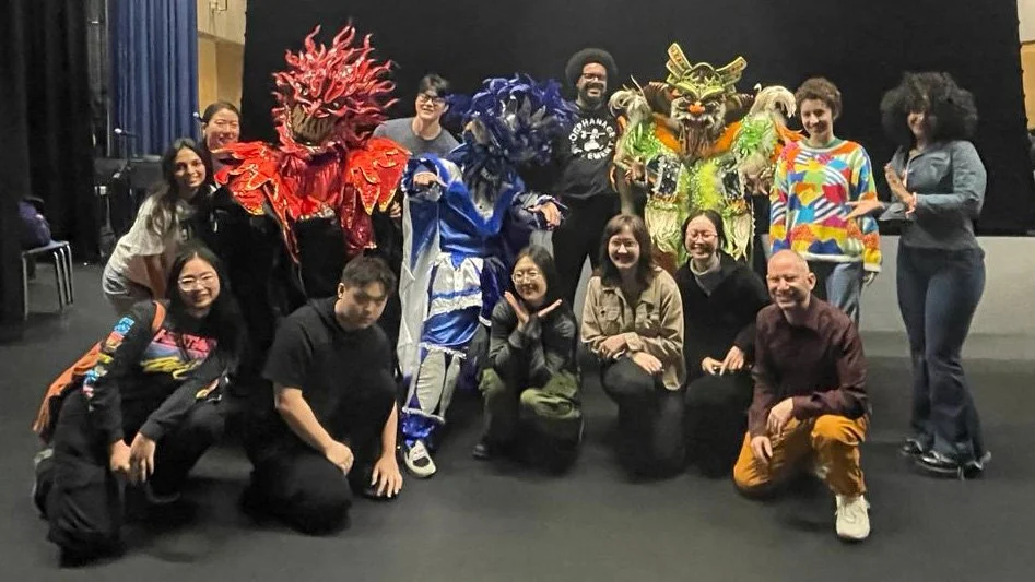 Group of people on stage with two elaborate traditional Asian lion dance costumes, one red and one yellow-green, and some in casual clothing.