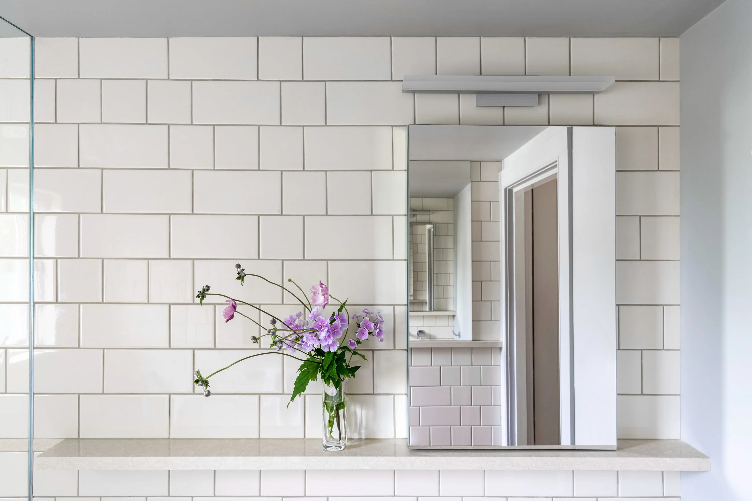 A bathroom mirror, wall-mounted light fixture, white subway-tiled wall, clear vase with purple flowers on white countertop.