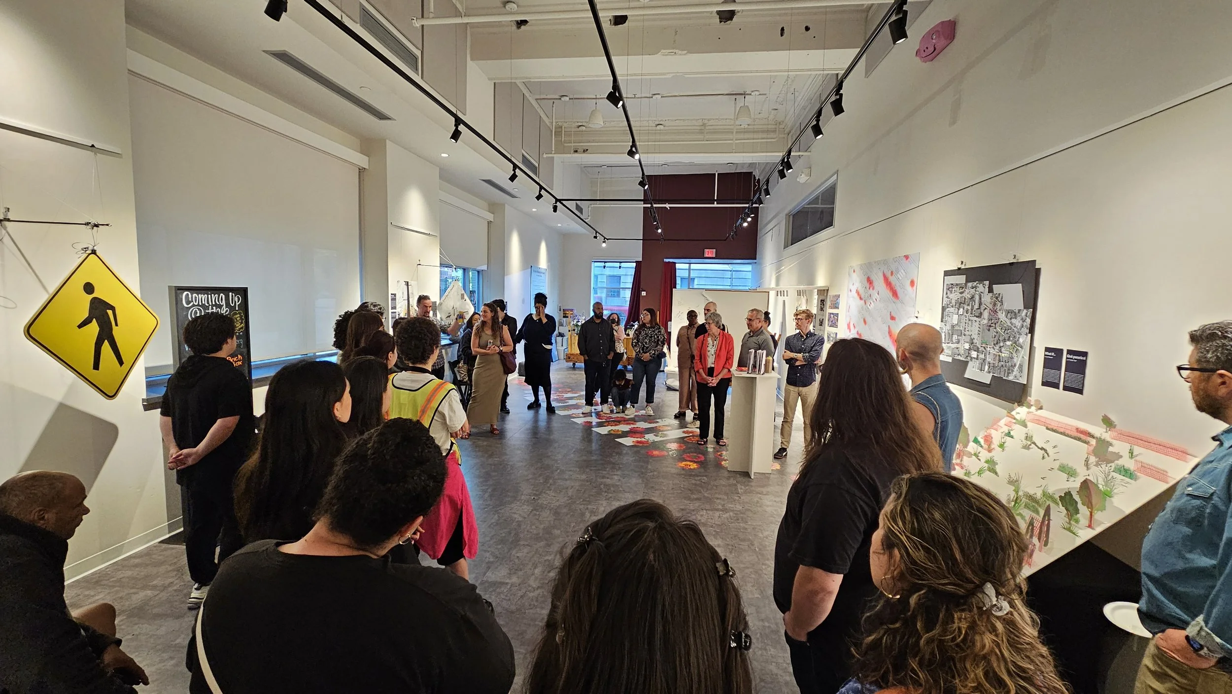 An art gallery with a diverse group of people gathered, looking at various art pieces and displays on the walls and floor. Some artwork features maps and abstract designs, and a yellow pedestrian crossing sign is visible on the left wall.