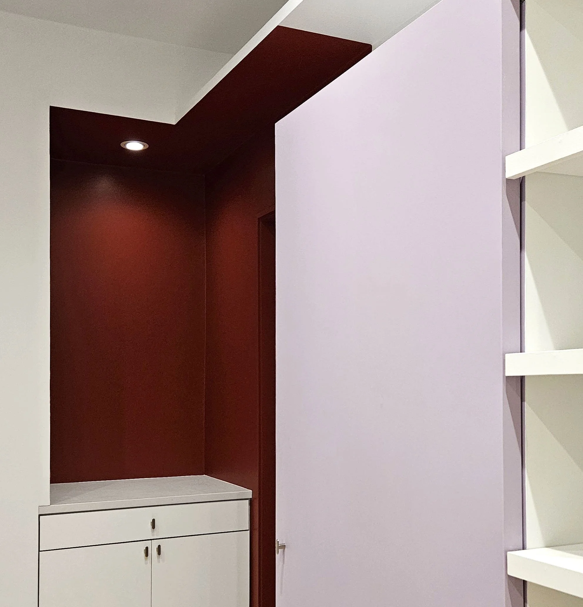 Interior of a modern room with a white and dark red wall, a white dresser with drawers, and a white shelf. Recessed ceiling light illuminates the space.
