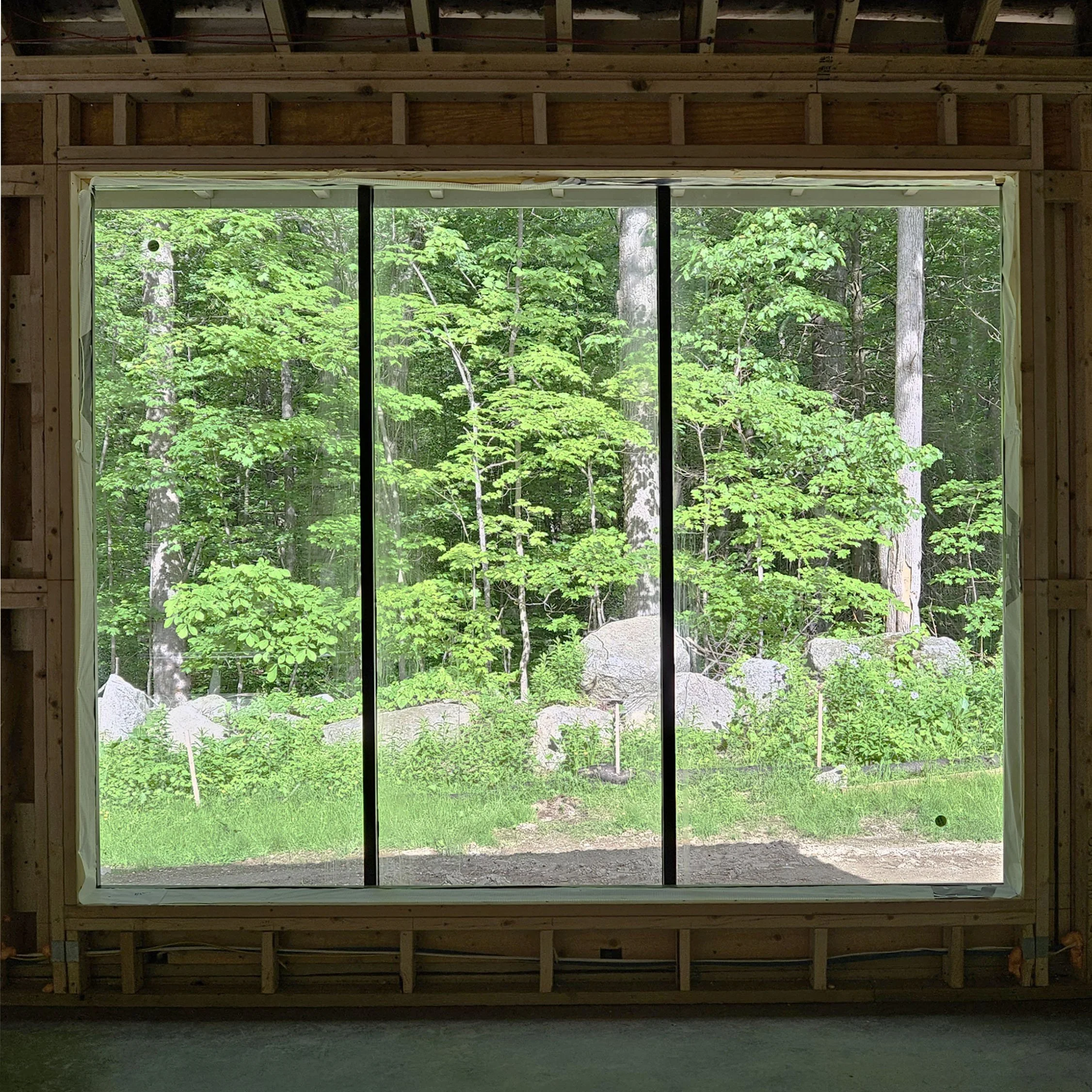 Unfinished wooden wall framing with a large window opening overlooking a green forest with trees and rocks.