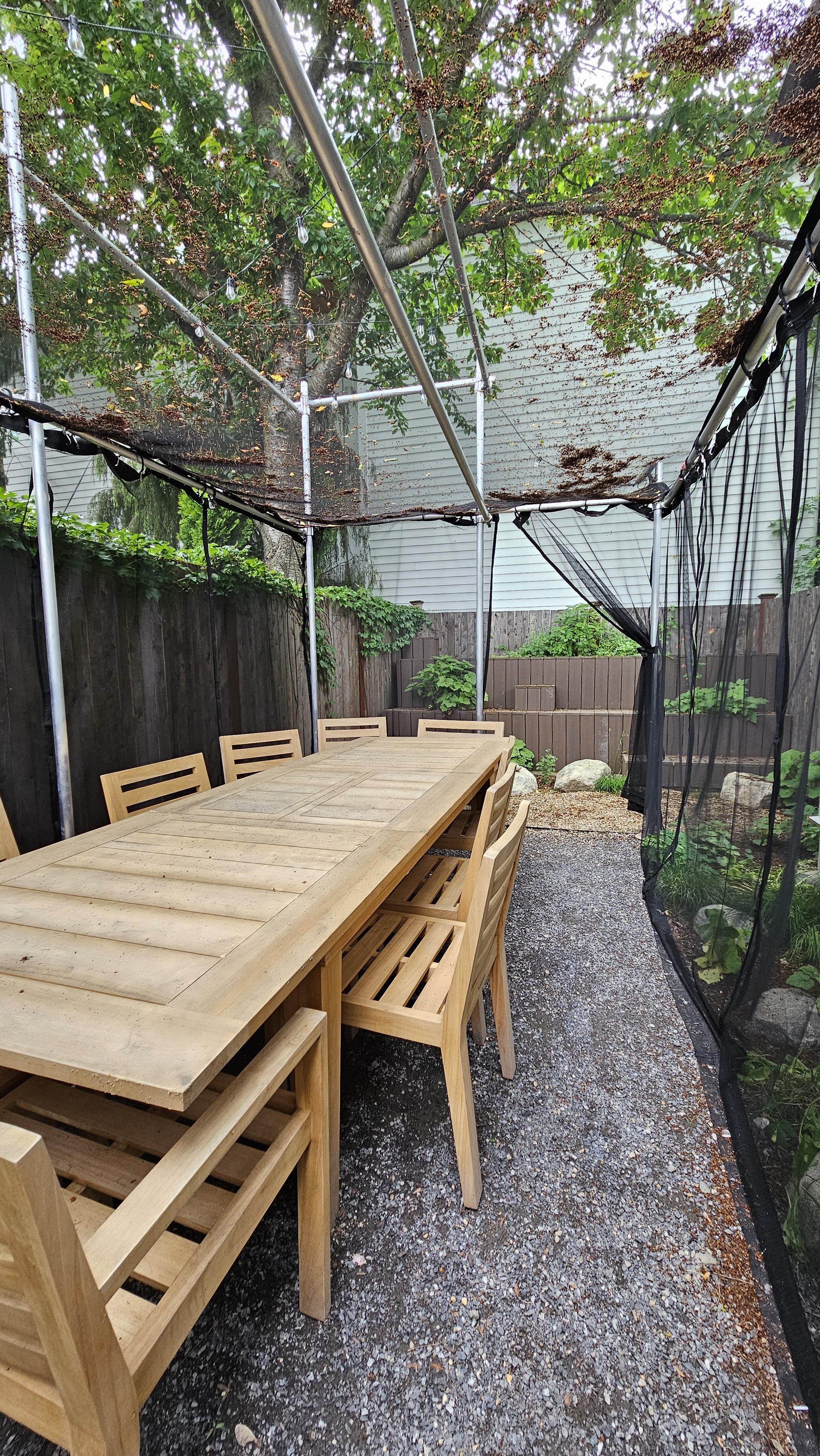An outdoor patio area with a long wooden dining table and multiple matching chairs, surrounded by a wooden fence, plants, and a gravel ground. A canopy with string lights overhead and a black mesh curtain on one side.