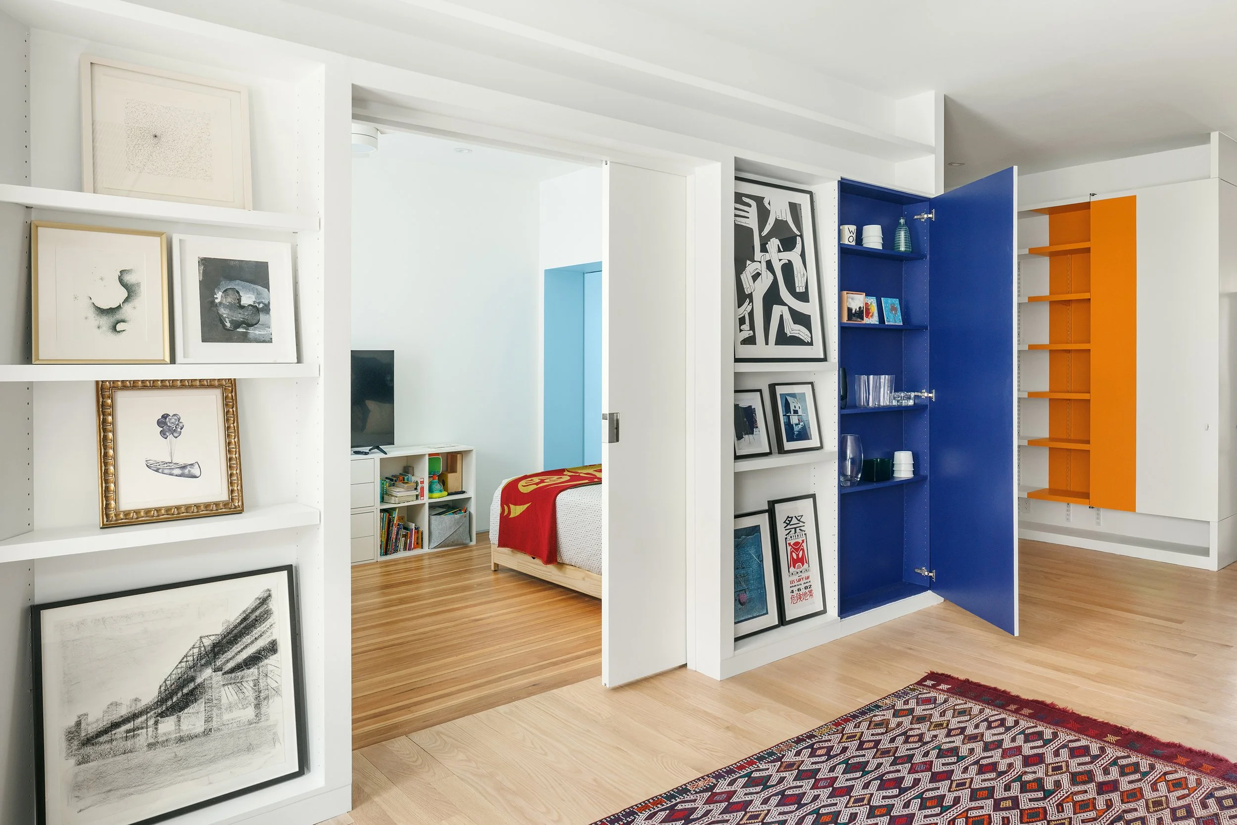 A modern, open-concept room featuring white walls and a decorative bookshelf with framed artwork, a colorful area rug, and a glimpse of a bedroom with a bed and books visible in the background.