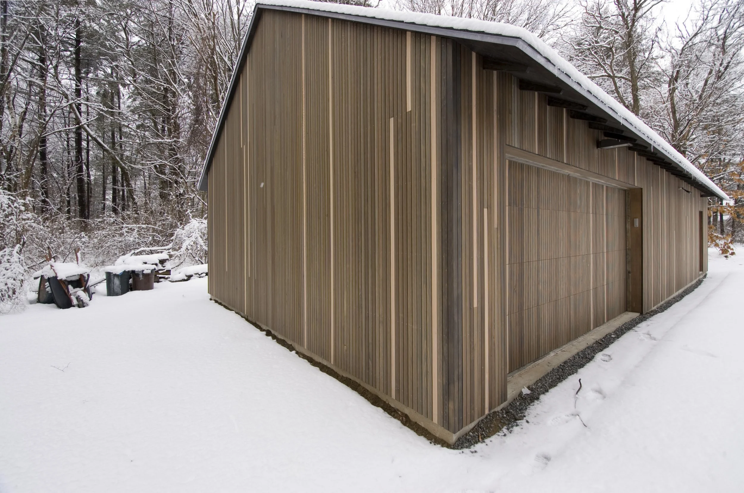 A modern wooden barn with vertical wood paneling situated in a snowy landscape surrounded by snow-covered trees.
