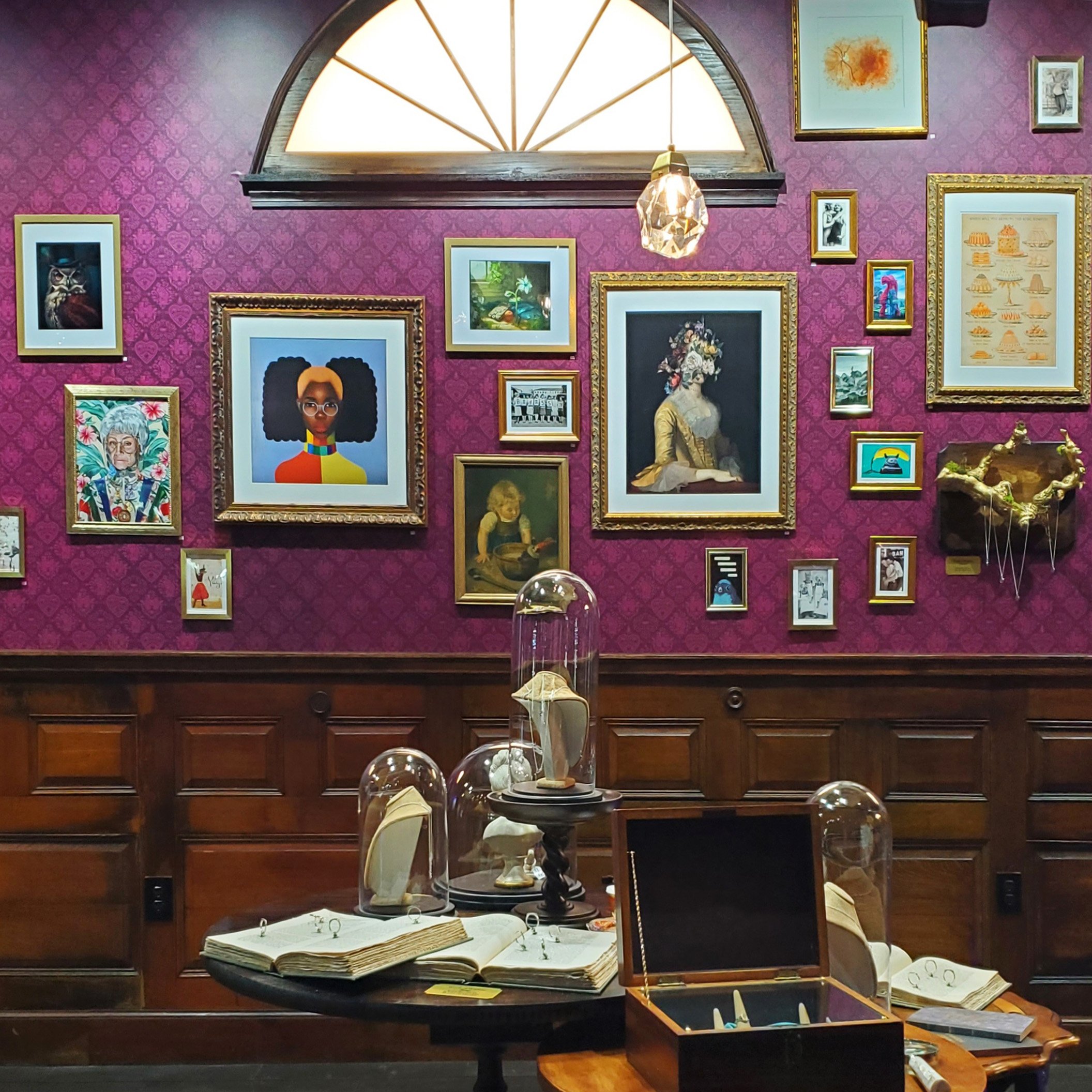 Display case with old books, jewelry, and small objects in a vintage shop or museum with framed pictures and artwork on a purple patterned wallpapered wall.