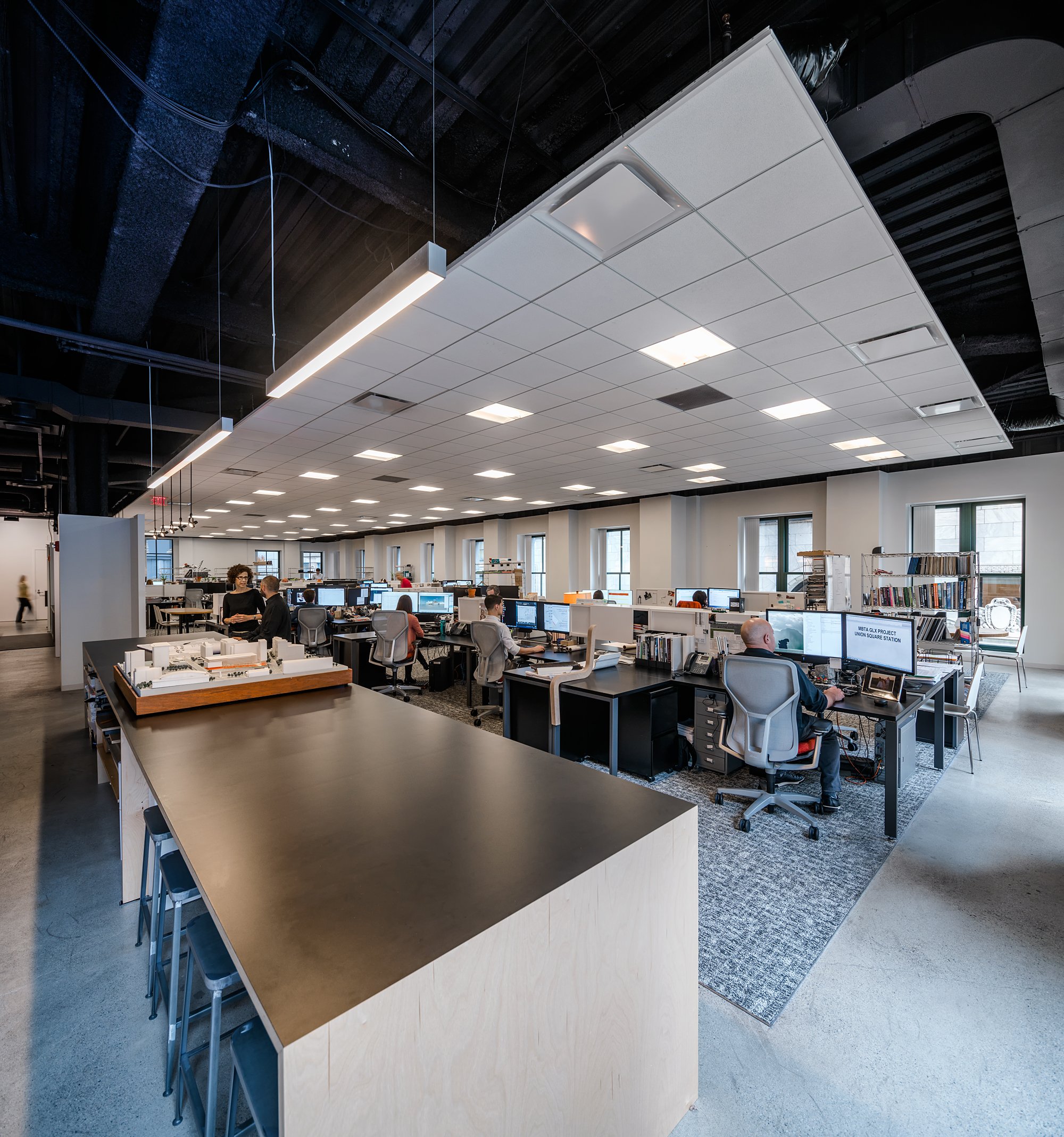 Open-plan office space with multiple workstations, computer monitors, and people working. Modern ceiling with suspended lights and a large table in the foreground.