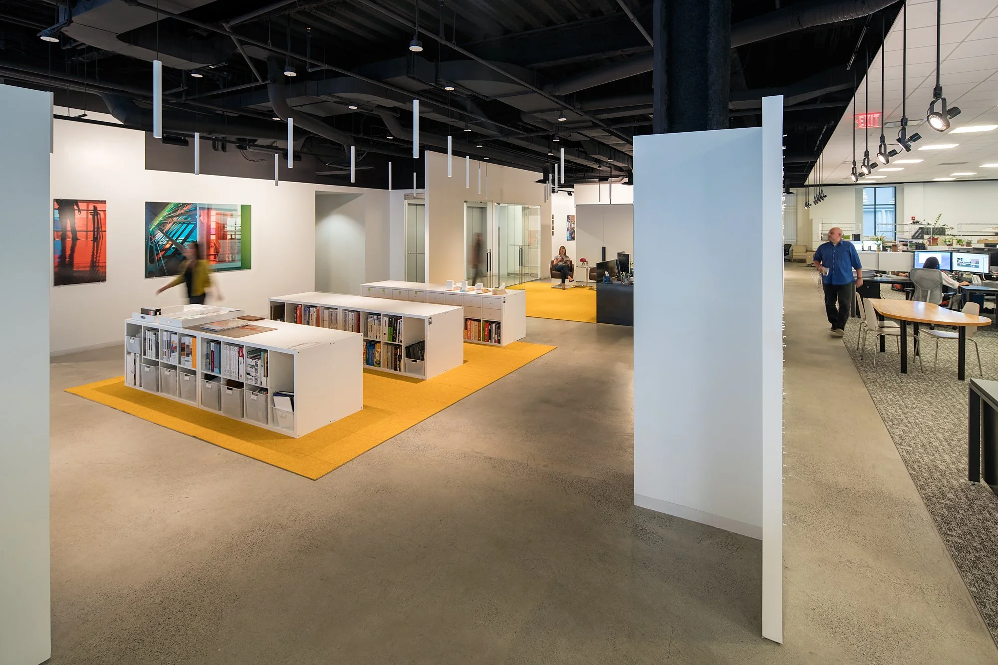 A modern office with white walls, artwork, and people working at desks. There are bookshelves, glass doors, and a yellow carpeted lounge area.
