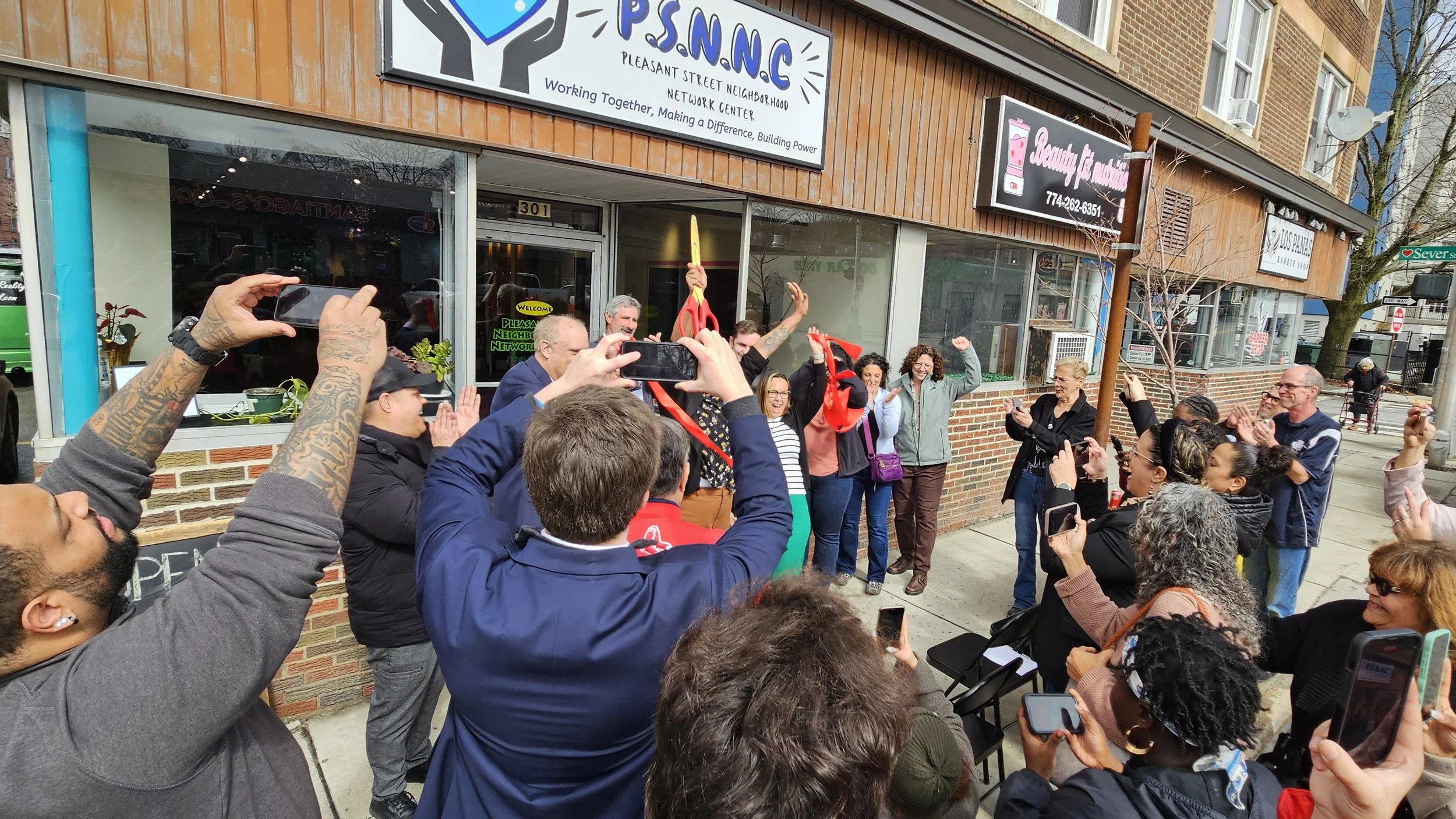 A diverse group of people gathered outside a building with a brick and wood facade, standing and taking photos of a woman cutting a red ribbon. The crowd is celebrating an event or opening, with some smiling and raising their hands.
