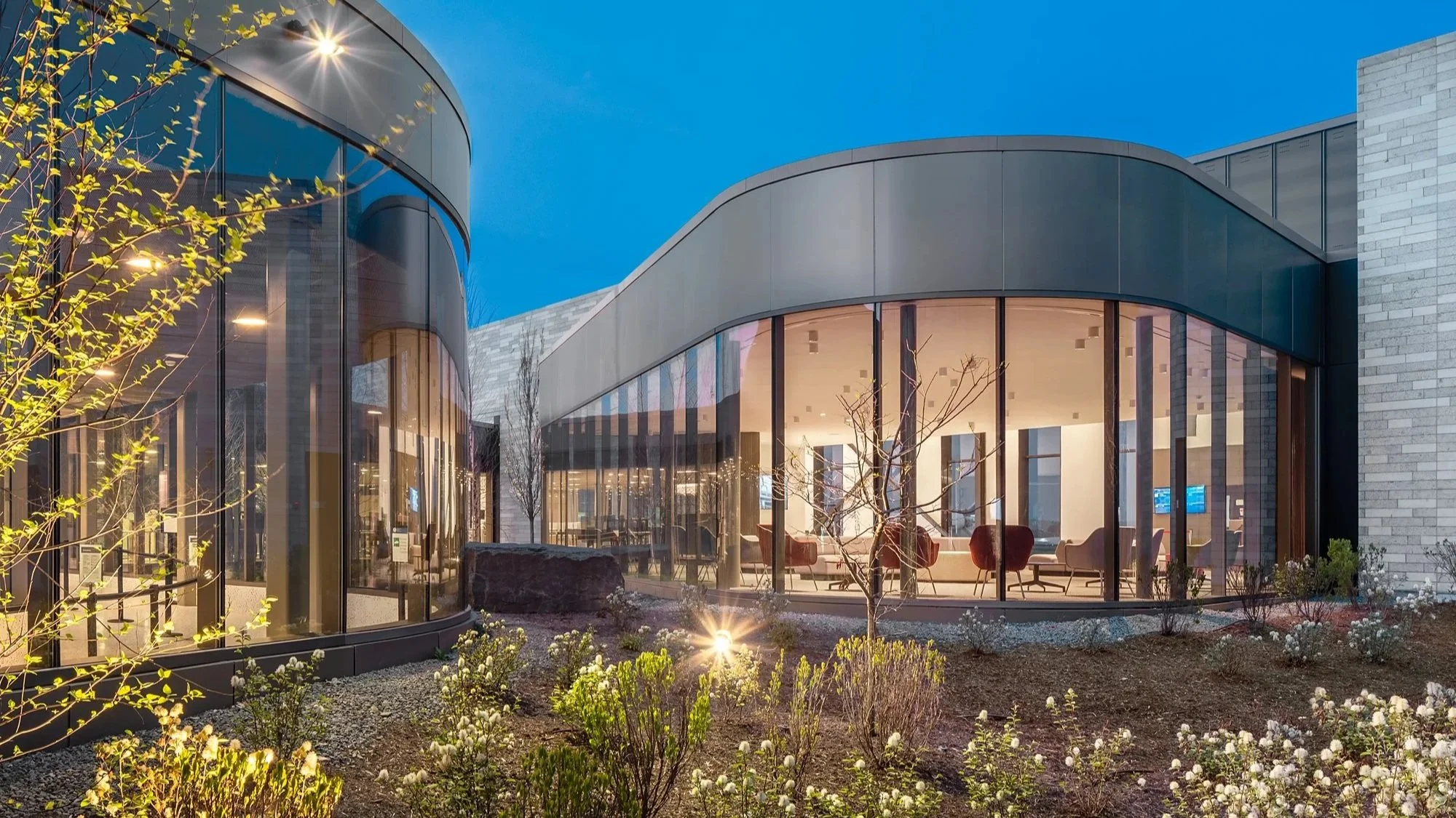 Airport with curved glass walls, surrounded by trees and landscaped garden, during dusk.