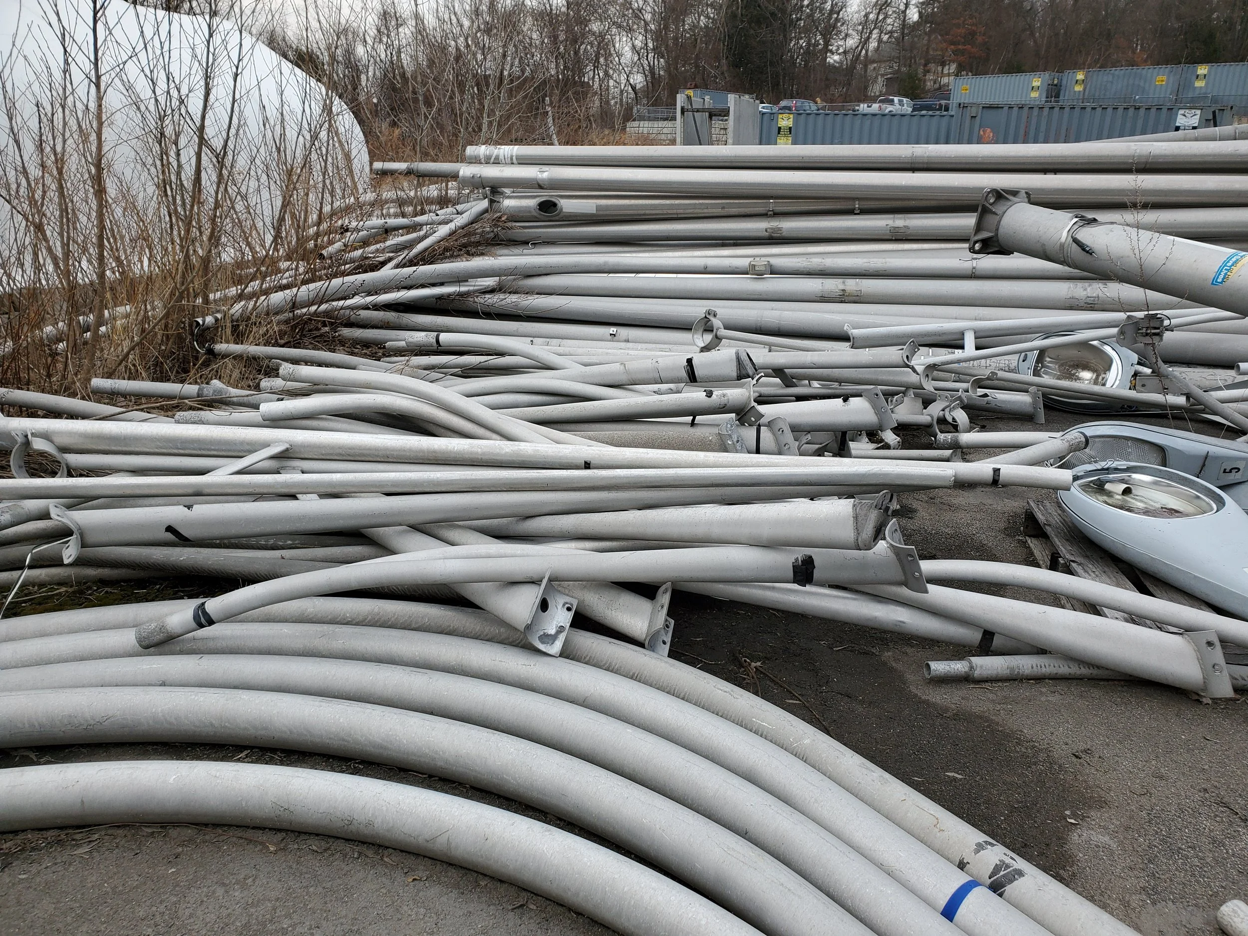 A pile of disorganized metal poles and pipes lying on the ground outdoors, with some among bushes and weeds, and shipping containers and parked cars in the background.