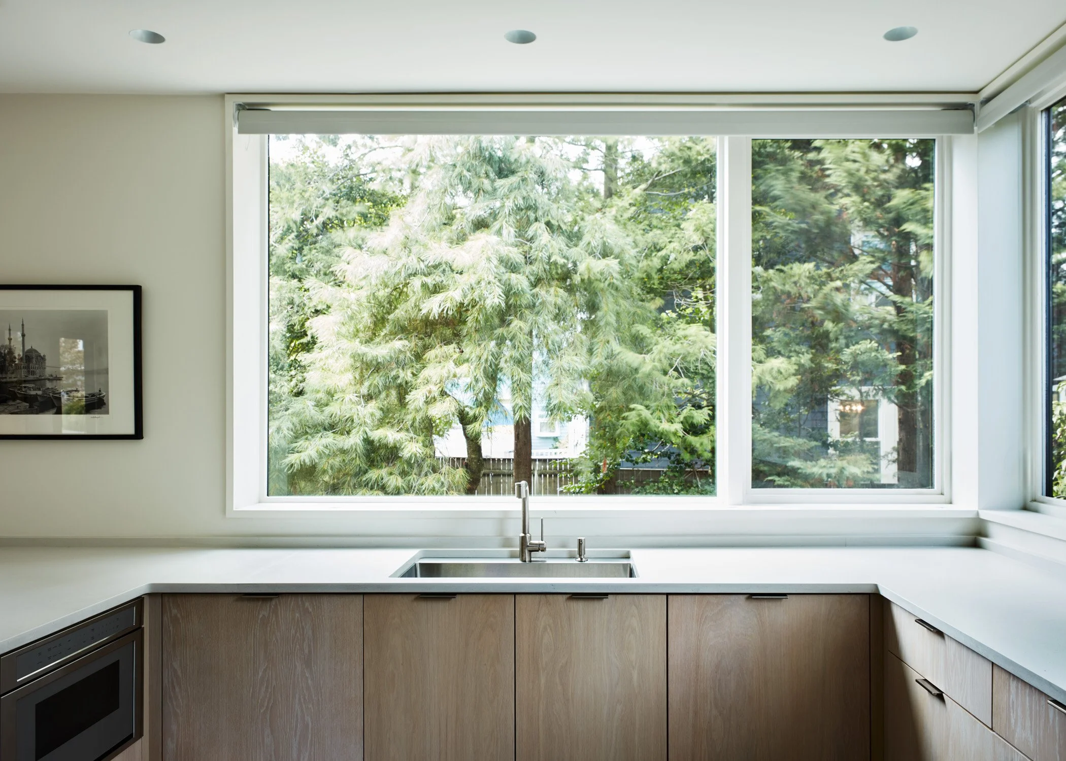 Modern kitchen with large window showing green trees outside, light-colored wood cabinets, a white countertop, and stainless steel sink.