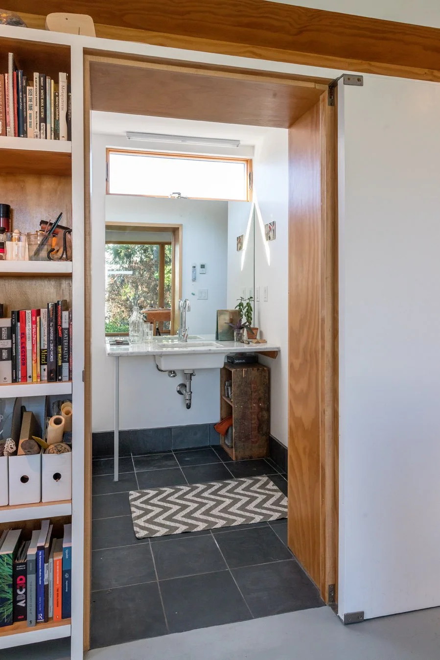 View through a doorway into a small bathroom with black floor tiles, a white sink, a small mirror, a window, and a potted plant on the counter.