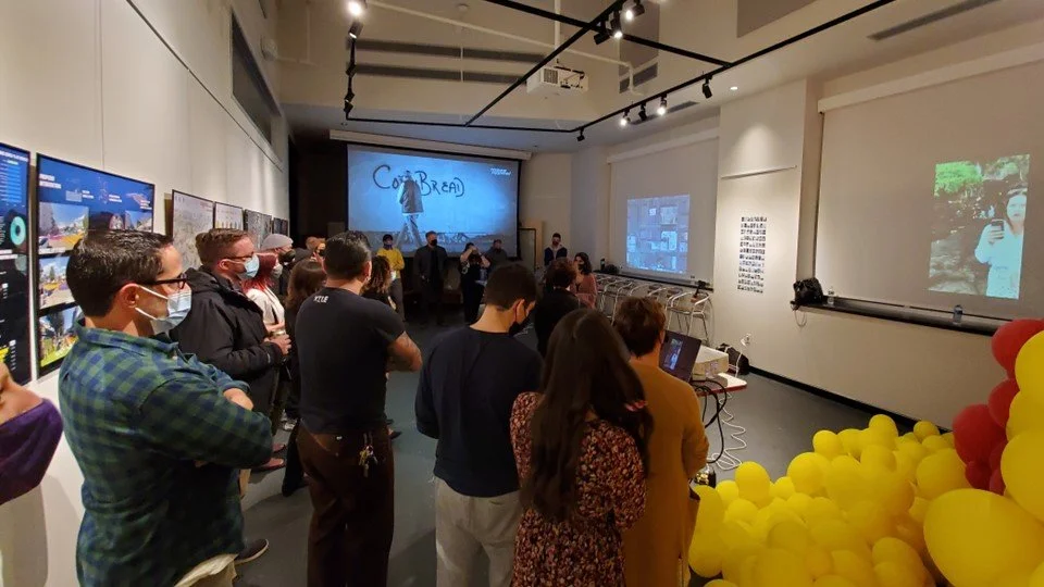 Group of people attending an indoor presentation or workshop, some wearing masks, with multiple monitors on the walls and a large screen at the front displaying a blackboard drawing and the word 'COLD BREAD'.