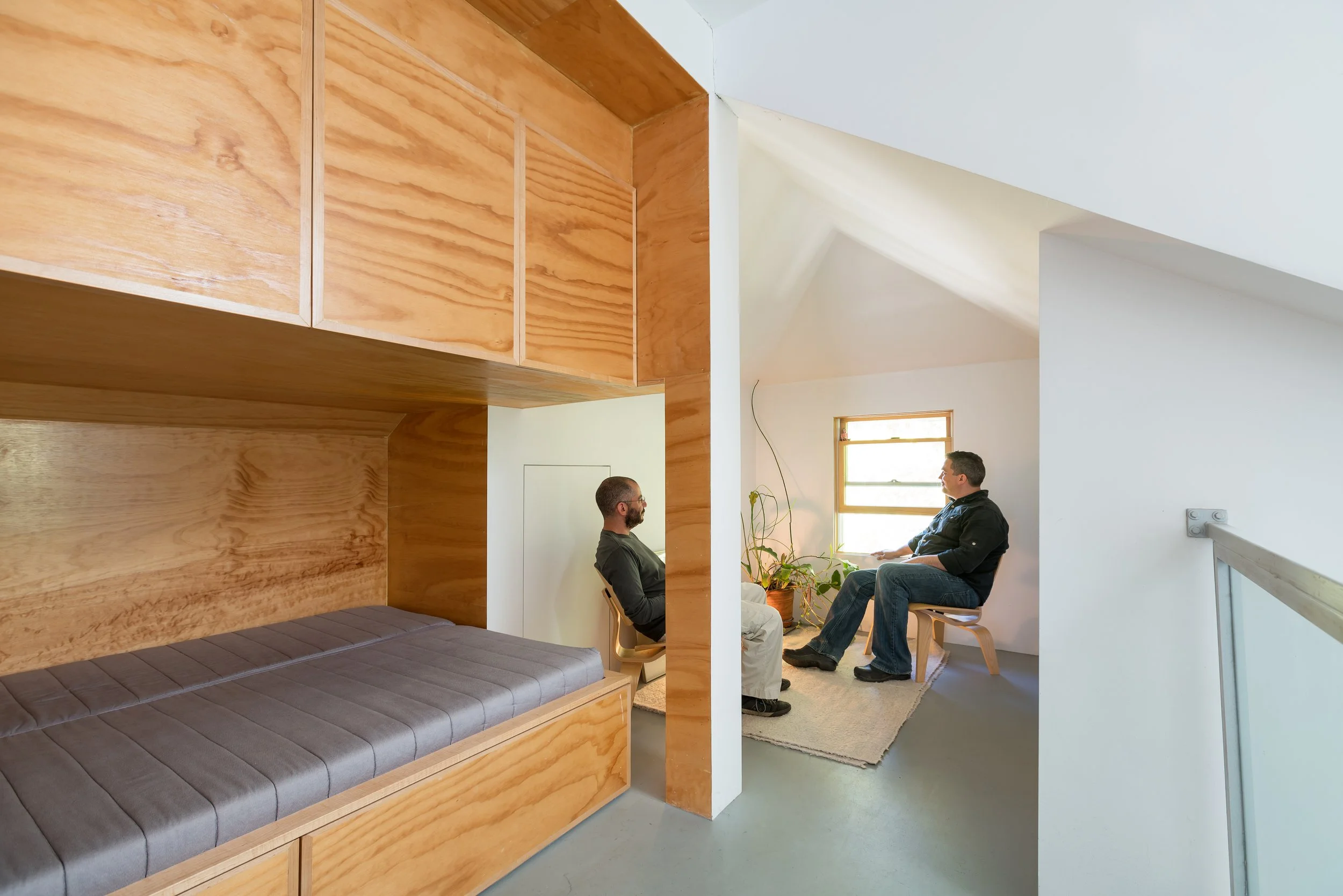 Two men sitting in chairs having a conversation in a bright, modern room with a window, a plant, and a wooden structure with built-in seating.