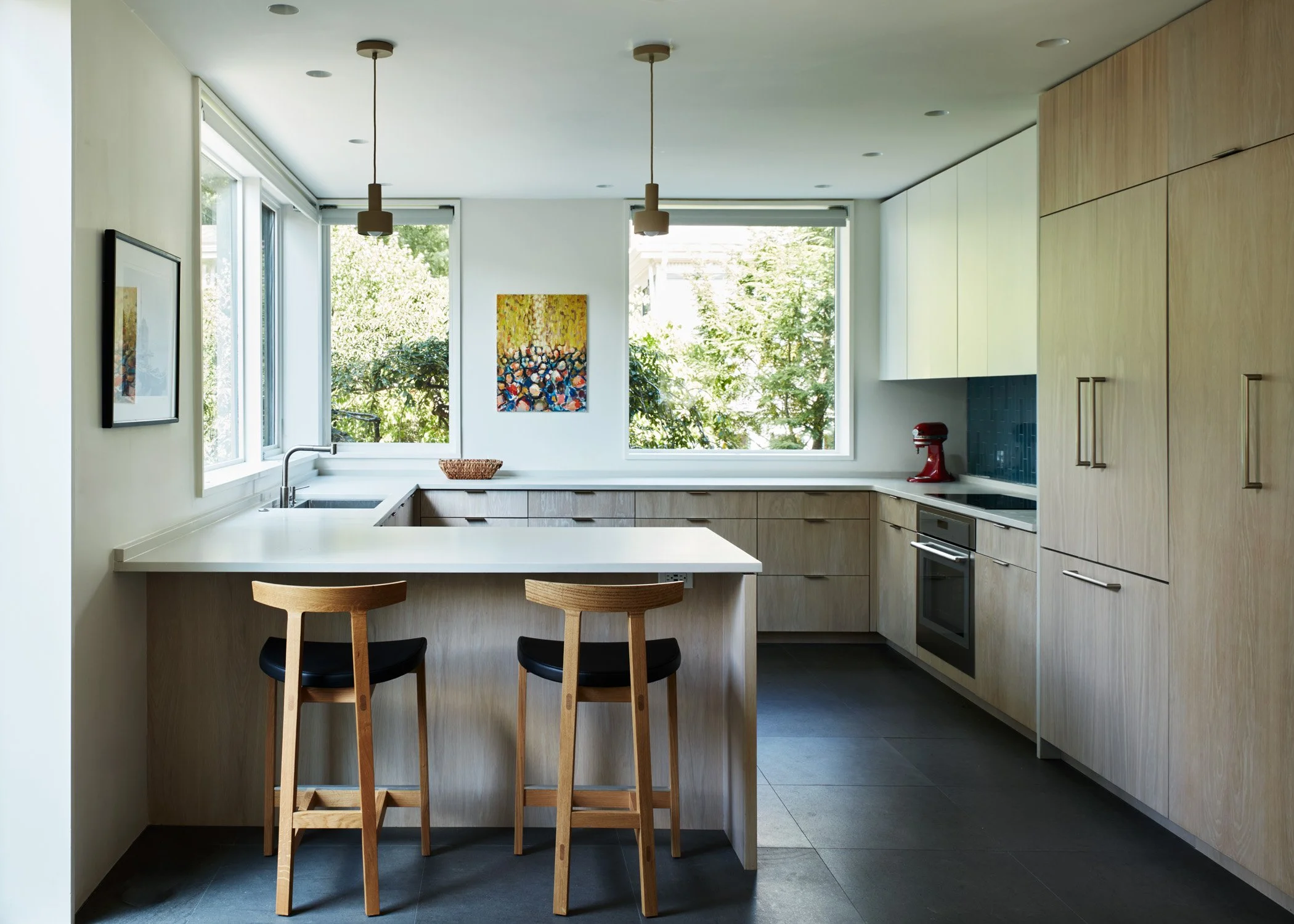 Modern kitchen with large windows, light wood cabinetry, black countertops, and two wooden barstools at a white kitchen island.
