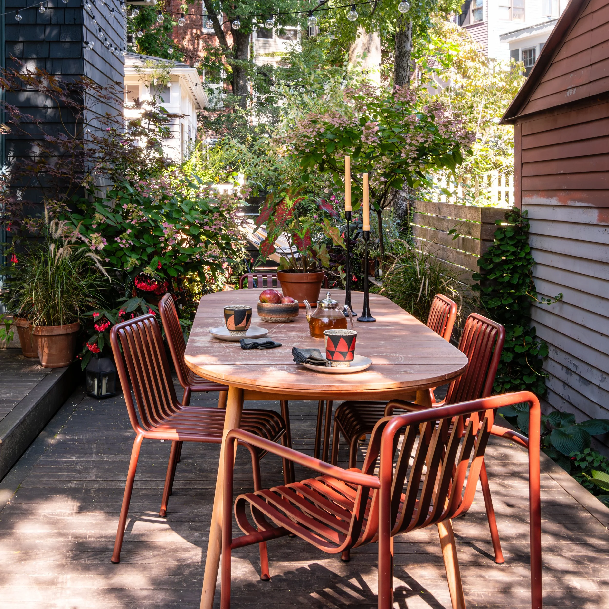 An outdoor patio with a wooden table set for a meal, surrounded by chairs and lush potted plants. String lights hang above, and there are trees and neighboring houses in the background.