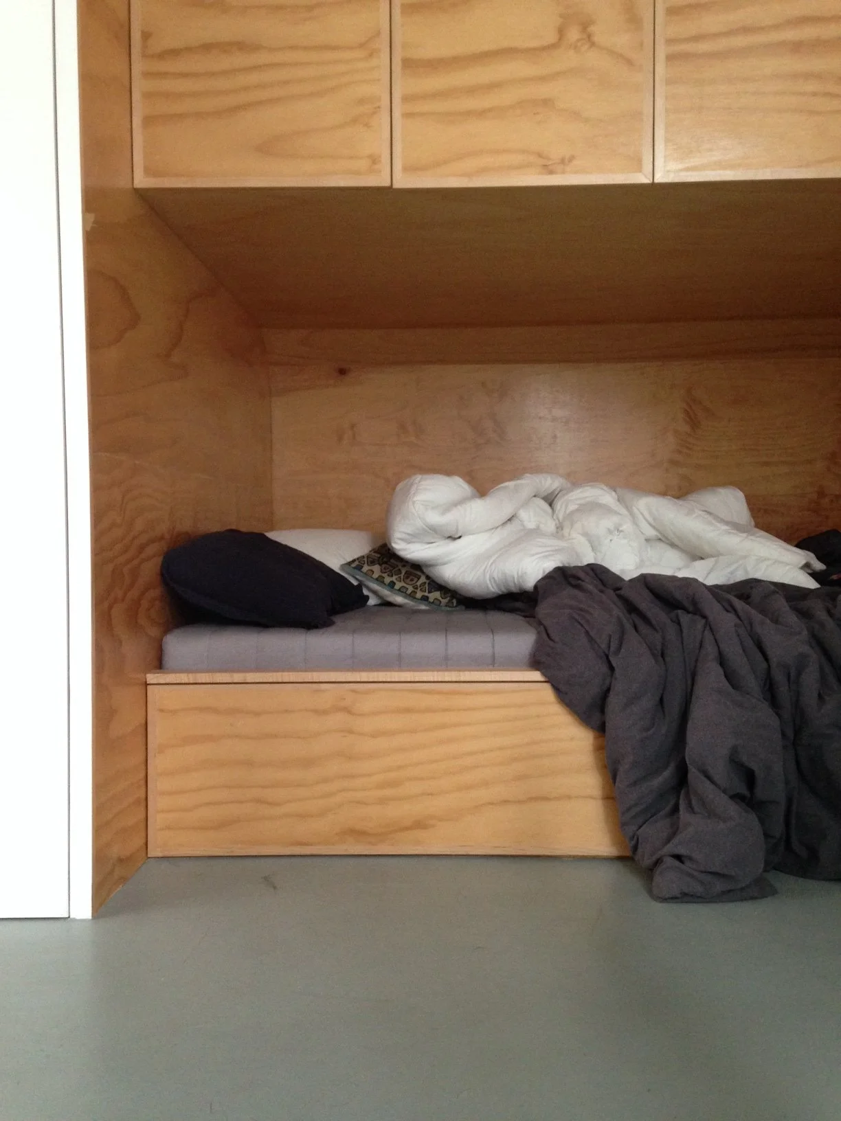 Unmade bed with pillows, blankets, and dark clothing on a wooden platform bed inside a room with wooden panel walls.