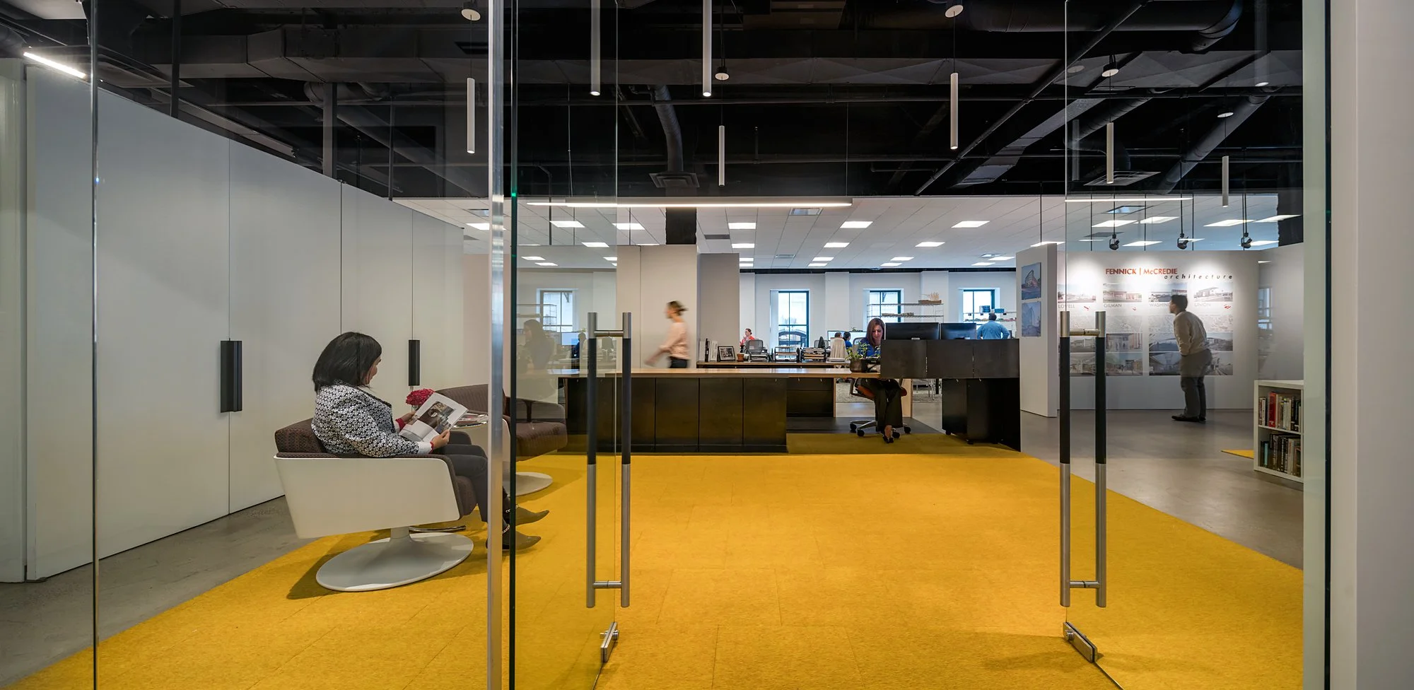 View of a modern office lobby with glass doors, yellow carpet, seating area, and people working inside.