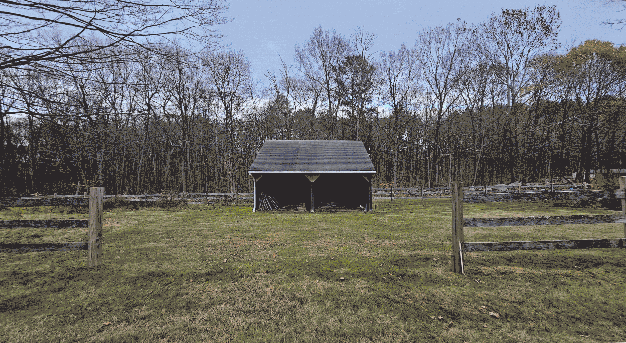 A black open-sided shed with a gabled roof on a grassy area, surrounded by a wooden fence and leafless trees in the background under a blue sky.