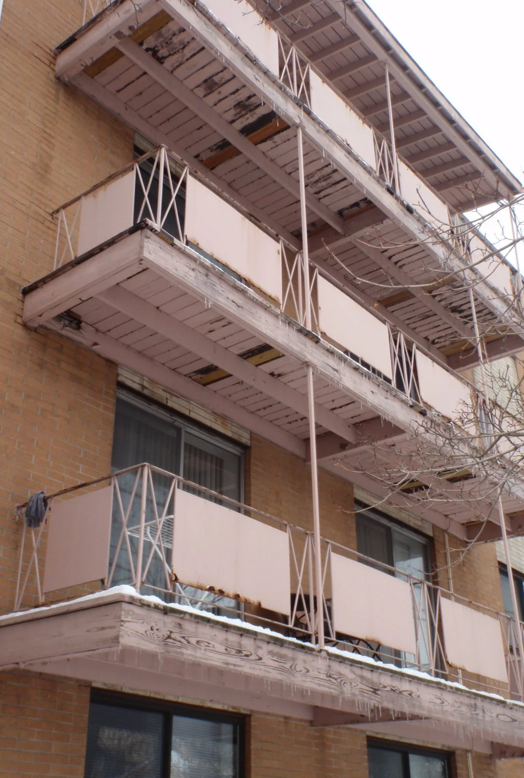 A brick apartment building with three balconies. The balconies appear worn, with peeling paint and some rust on the metal railings.