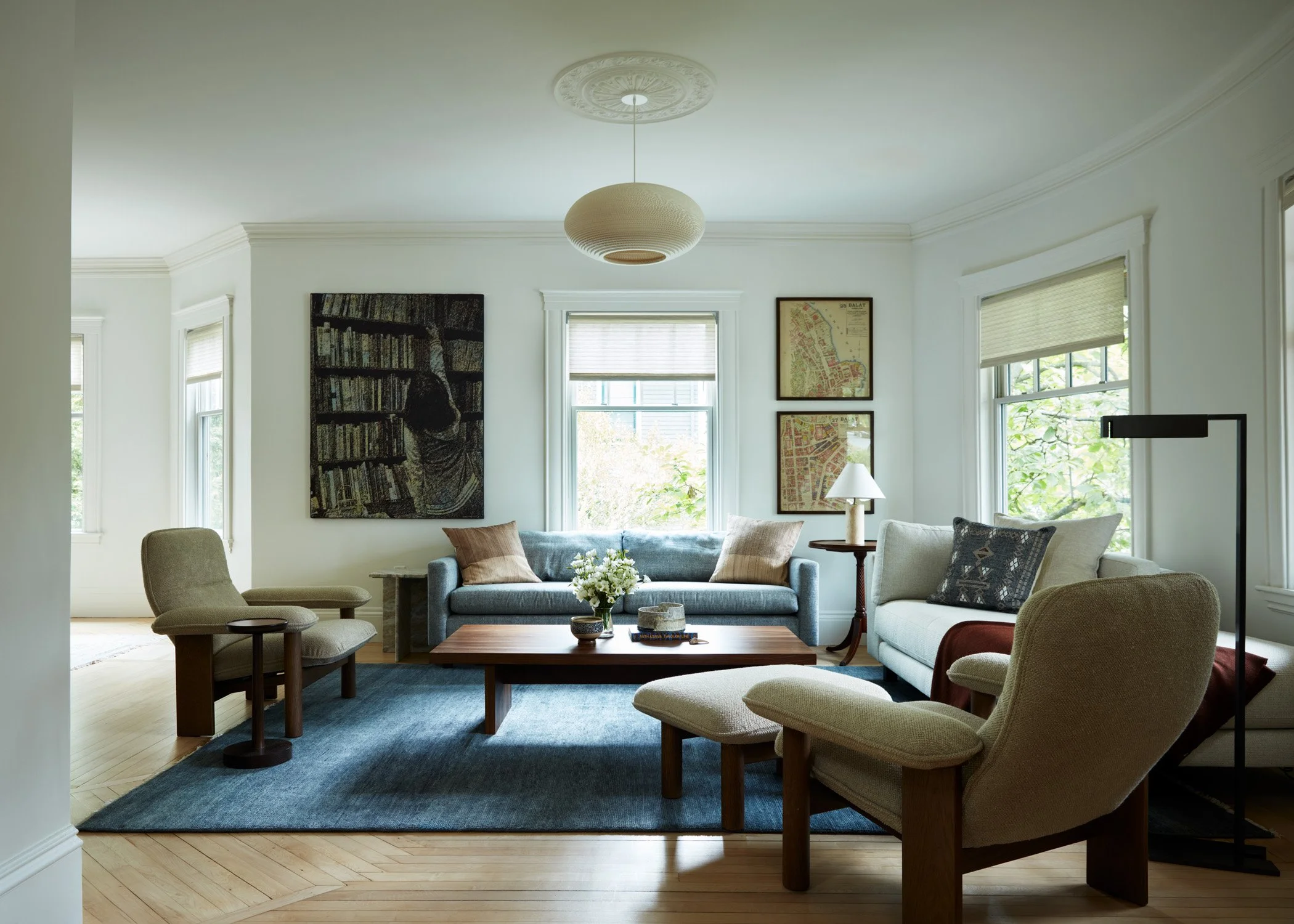 A modern living room with large windows, white walls, a gray sofa, beige chairs, a wooden coffee table with books and a flower arrangement, framed artwork on the walls, a white lampshade, and a blue rug.