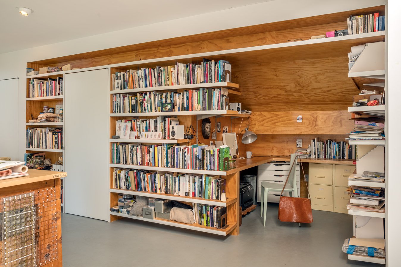 A home office with wooden built-in shelves filled with books, a desk with a chair, and various office supplies, against a wooden accent wall.