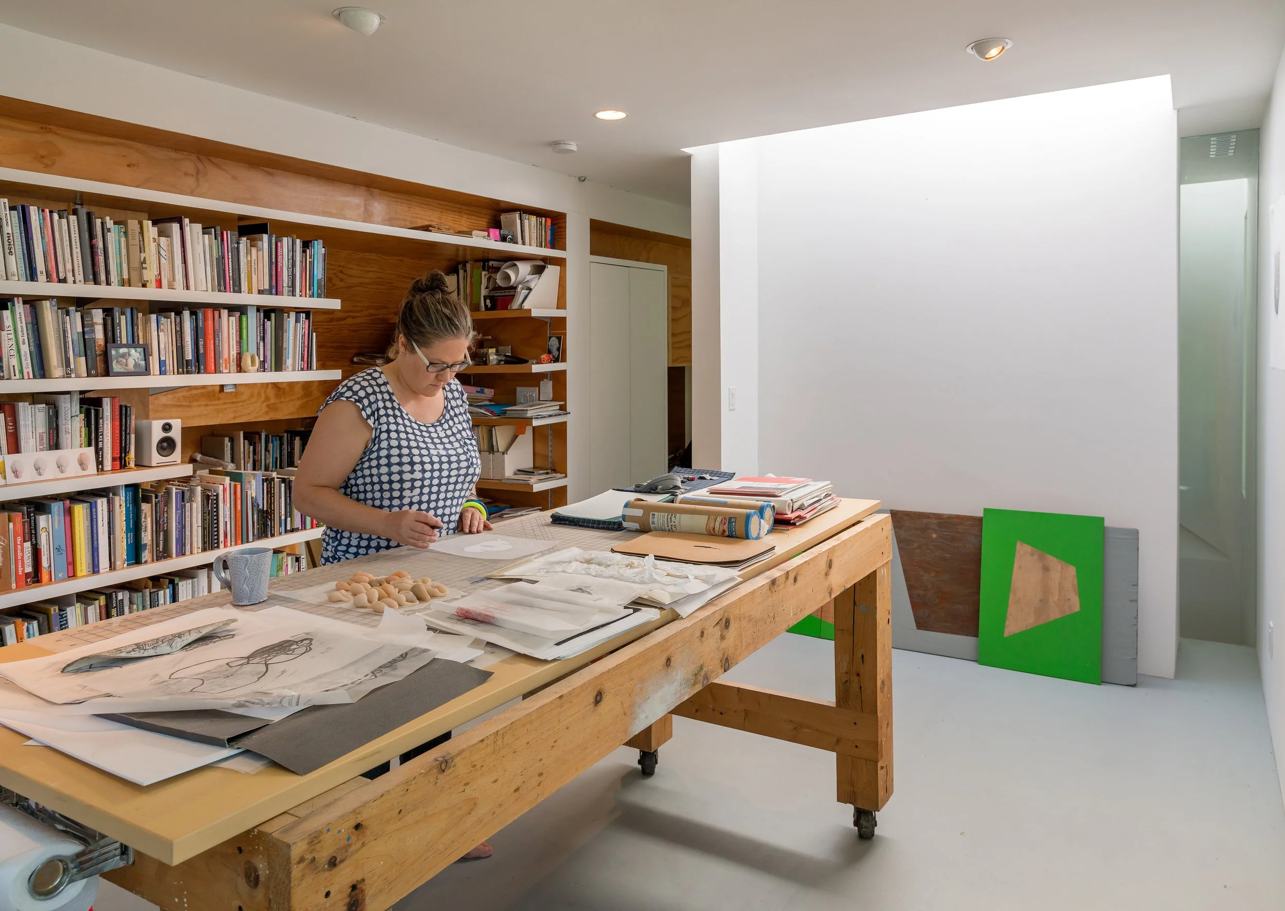 A woman working at a large wooden table in an art studio or library, with a computer and stacks of papers, books, and art supplies. Behind her, white bookshelves filled with books and a modern white wall with green and brown panels.