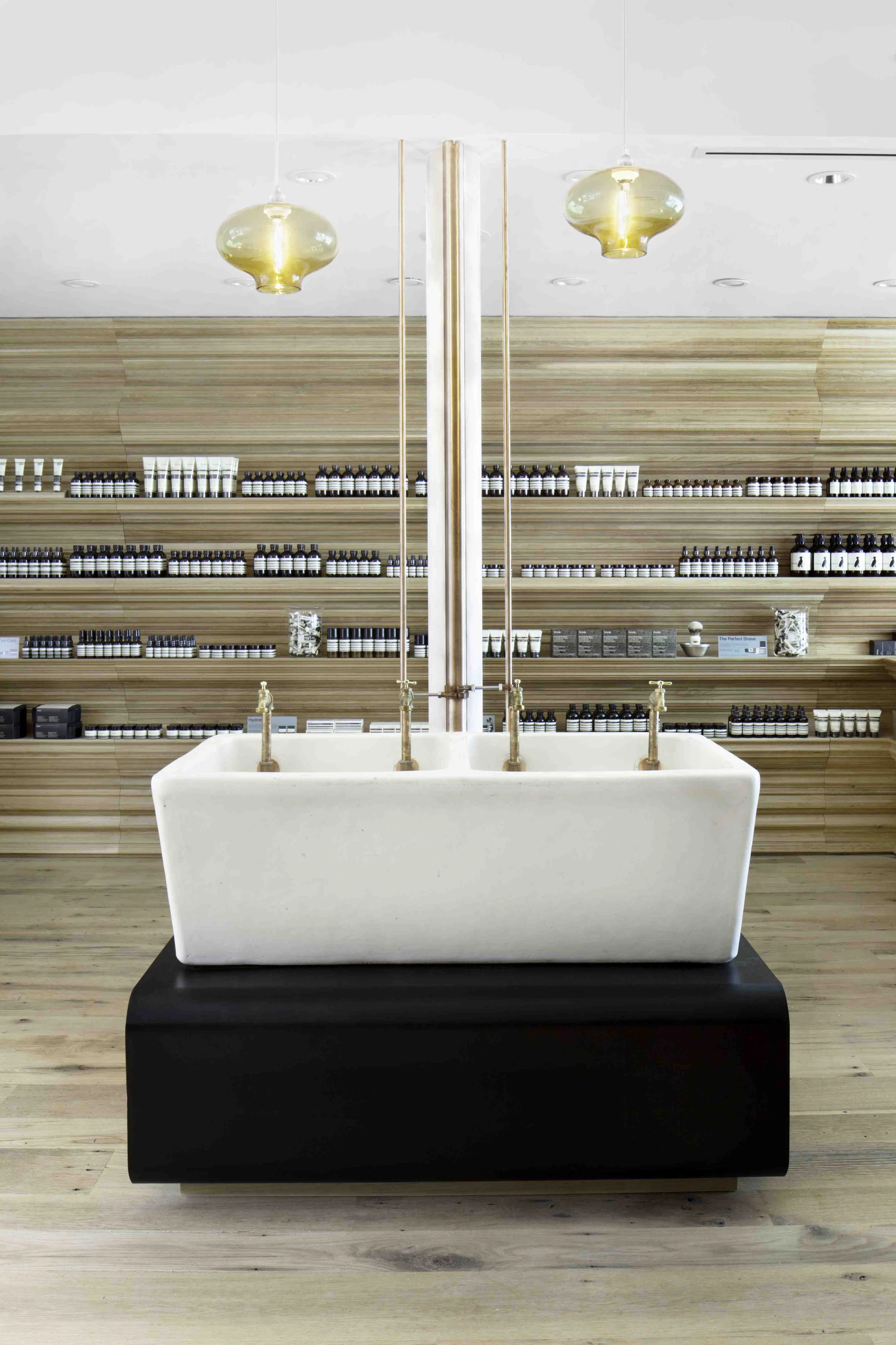 A modern storefront with a large white bathtub on a black platform, copper fixtures, and a wooden wall with shelves filled with small bottles and skincare products, illuminated by two glass pendant lights.