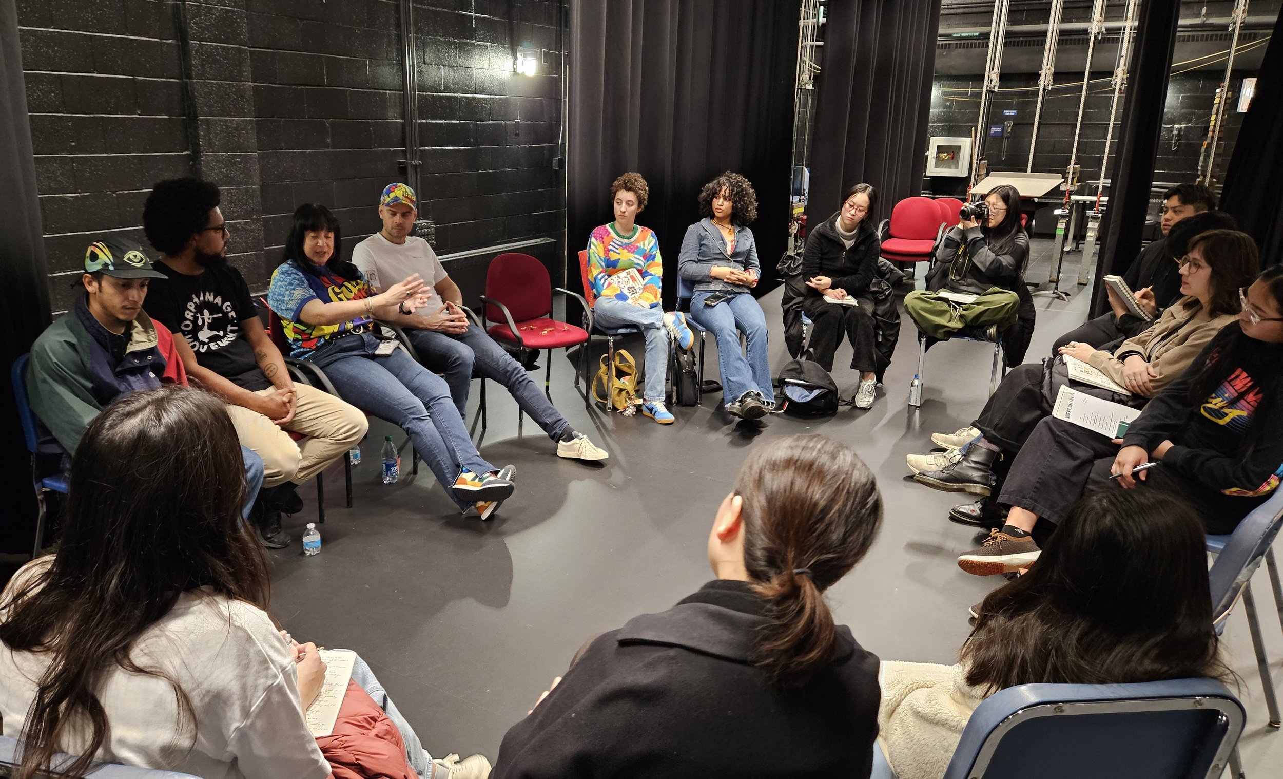A diverse group of people sitting in a circle in a black-walled room, engaged in a discussion or meeting, some taking notes or holding books.