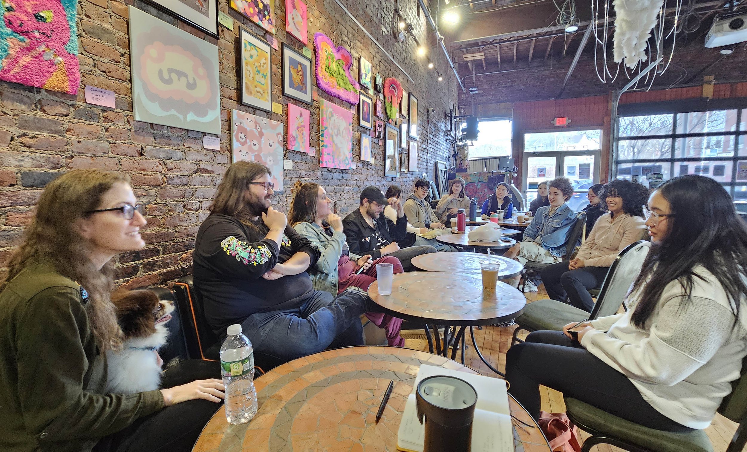A group of students sitting in a cozy art gallery with a brick wall decorated with colorful artwork. The group is engaged in discussion.