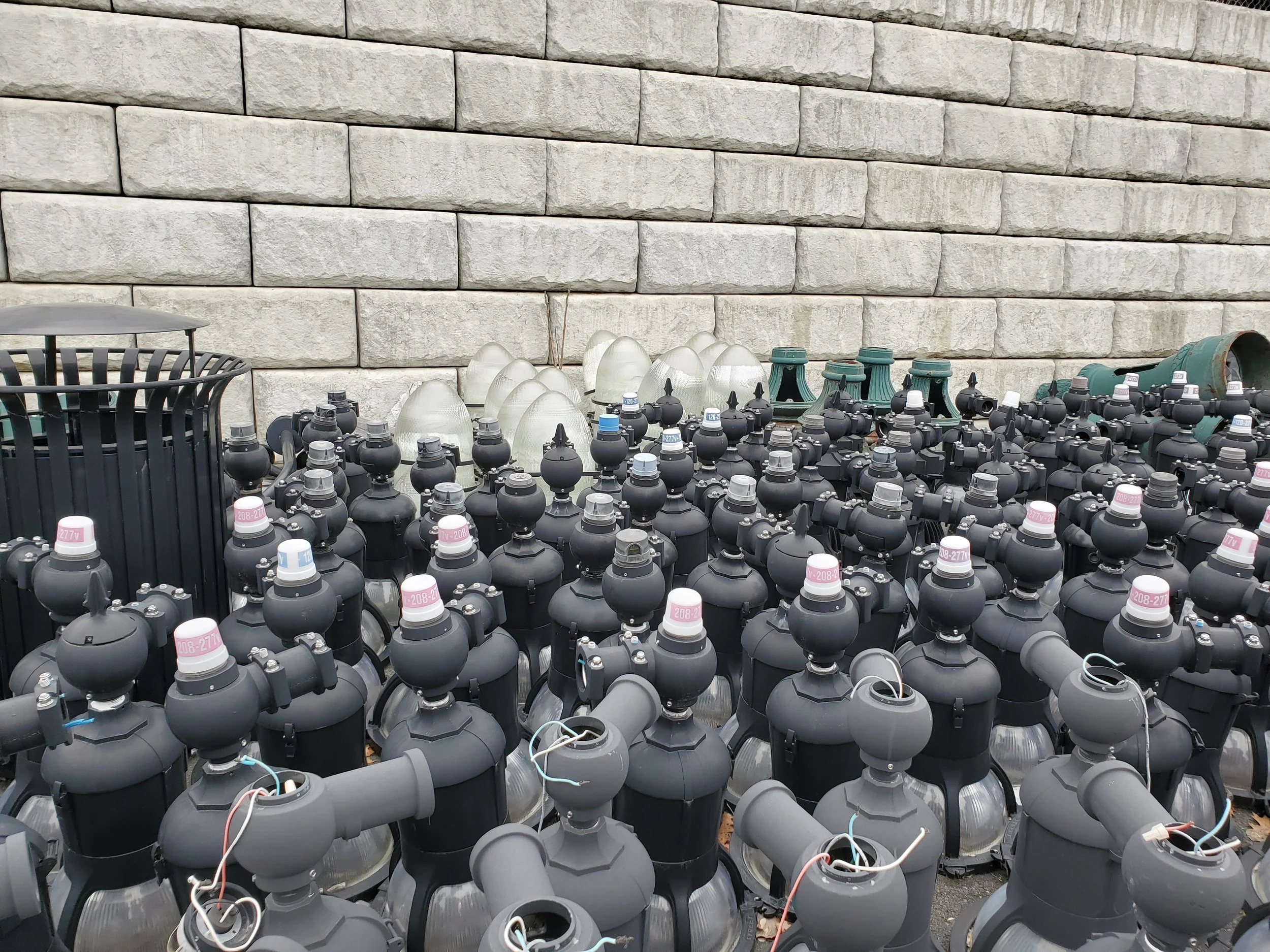 An outdoor area with many used LED streetlight fixtures, some with wires exposed, laid out on the ground against a stone wall.