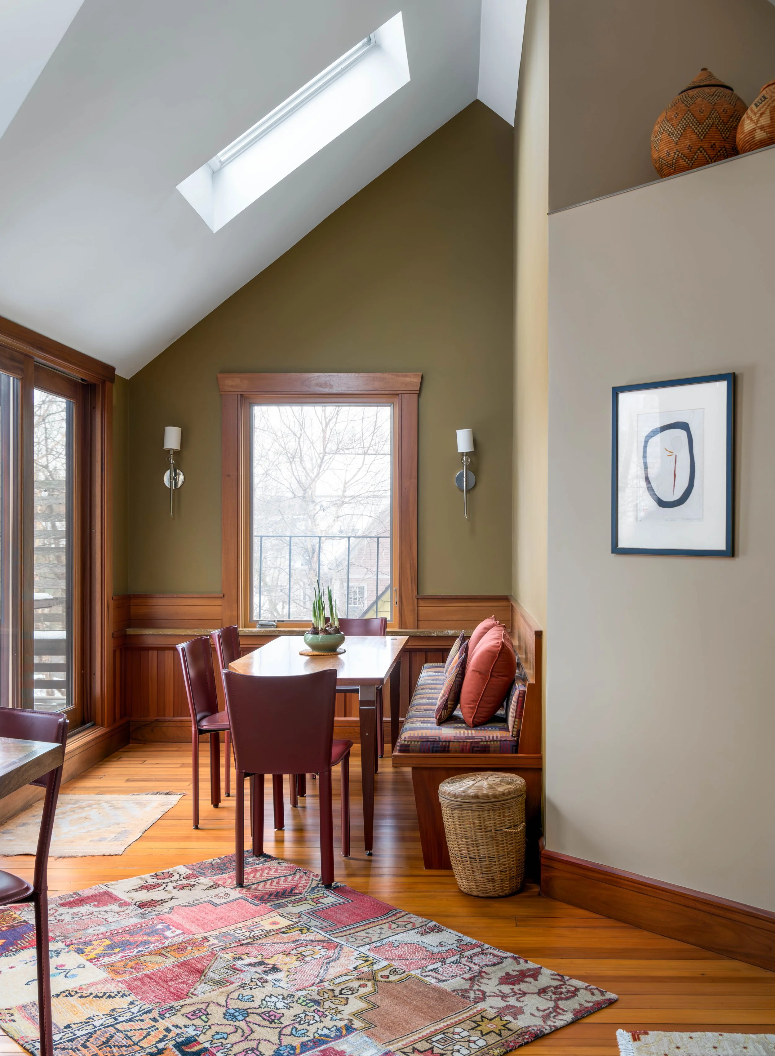 Interior view of a dining room with hardwood floors, a wooden dining table, and red chairs. There's a built-in corner bench with patterned cushions and a window with natural light coming through. A decorative rug is on the floor, and a pair of wall-m