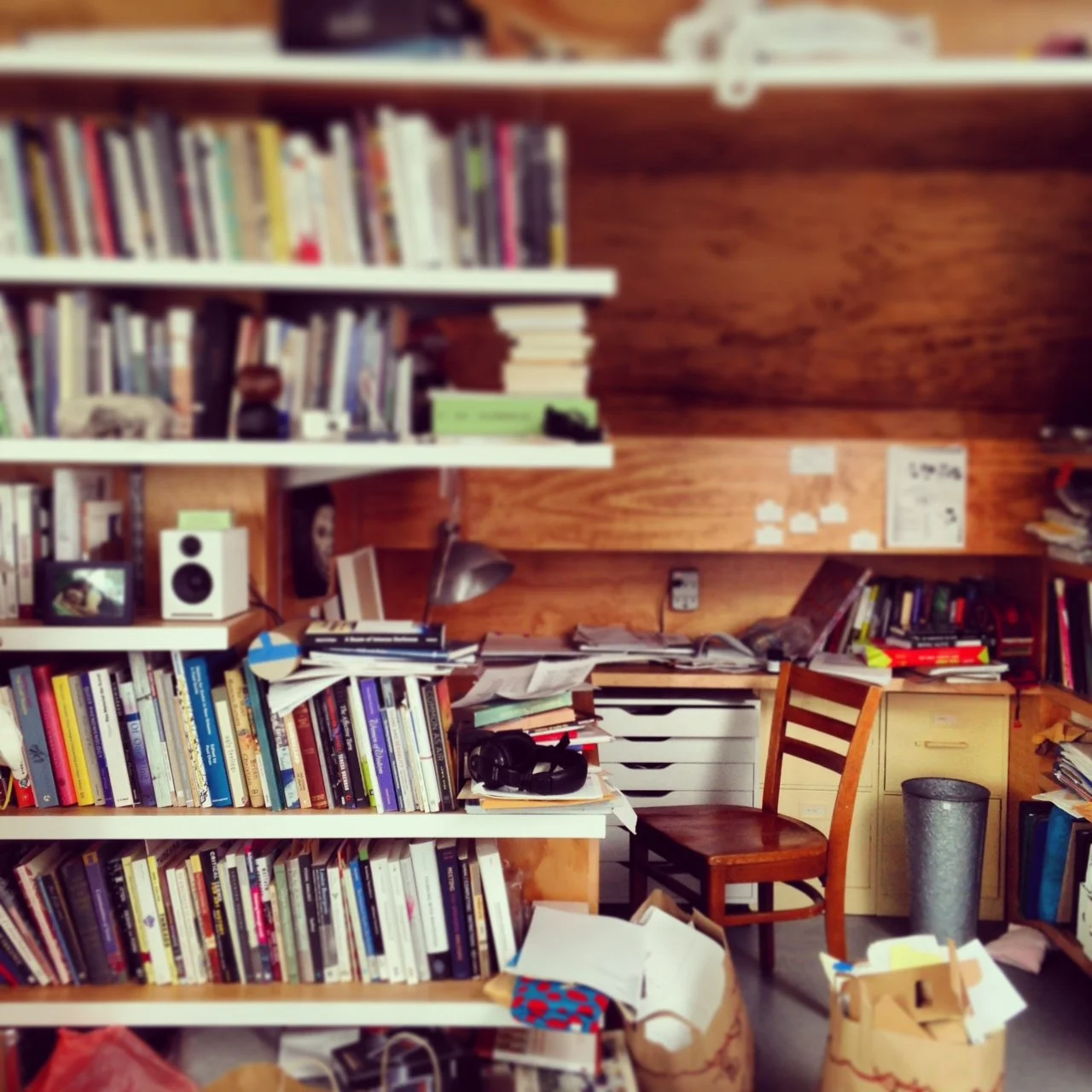 A cluttered home office or study with bookshelves filled with many books, a wooden desk with various papers, a chair, and a trash bin.