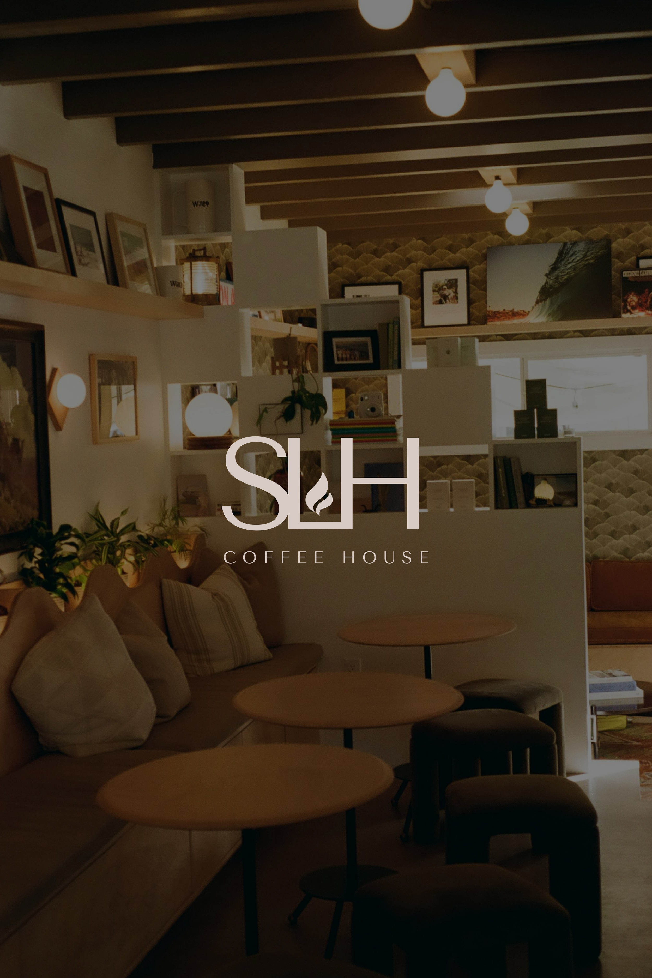 Interior of a cozy coffee shop with a beige sofa, cushions, round wooden tables, and black stools. The space is decorated with framed pictures, plants, and warm, warm lighting from ceiling light bulbs. The logo 'USH COFFEE HOUSE' is visible at the bottom of the image.