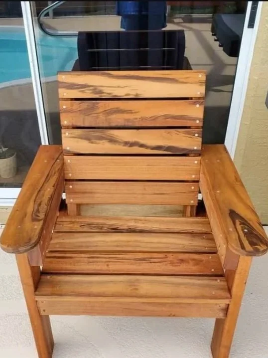 A wooden outdoor chair with wide armrests and slatted backrest and seat, positioned on a patio or balcony with a glass door and pool in the background.