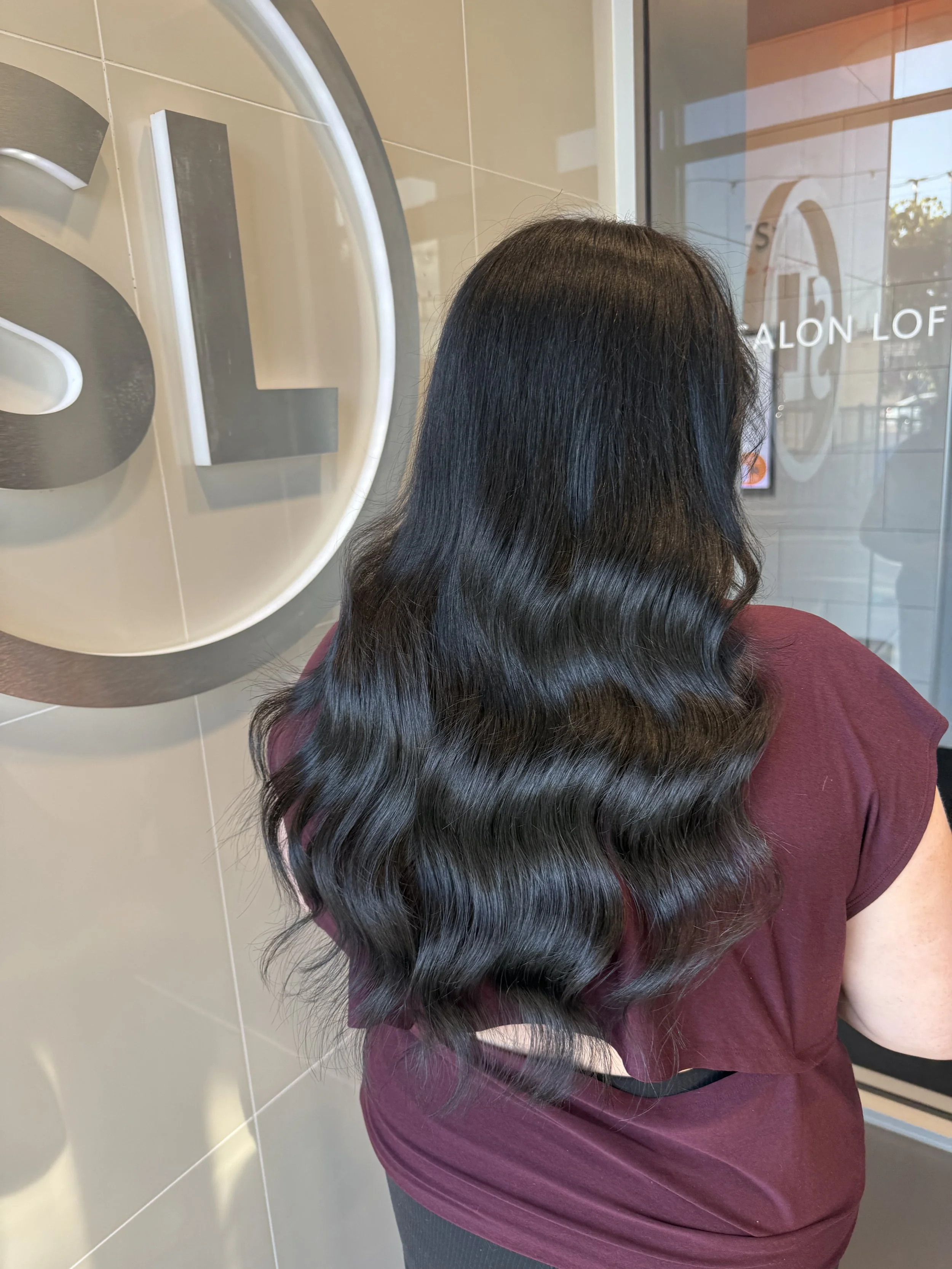 Woman with long, dark, wavy hair extensions , wearing a maroon top, standing beside a mirror with a salon logo, in a salon waiting area with a glass door and tiled wall.