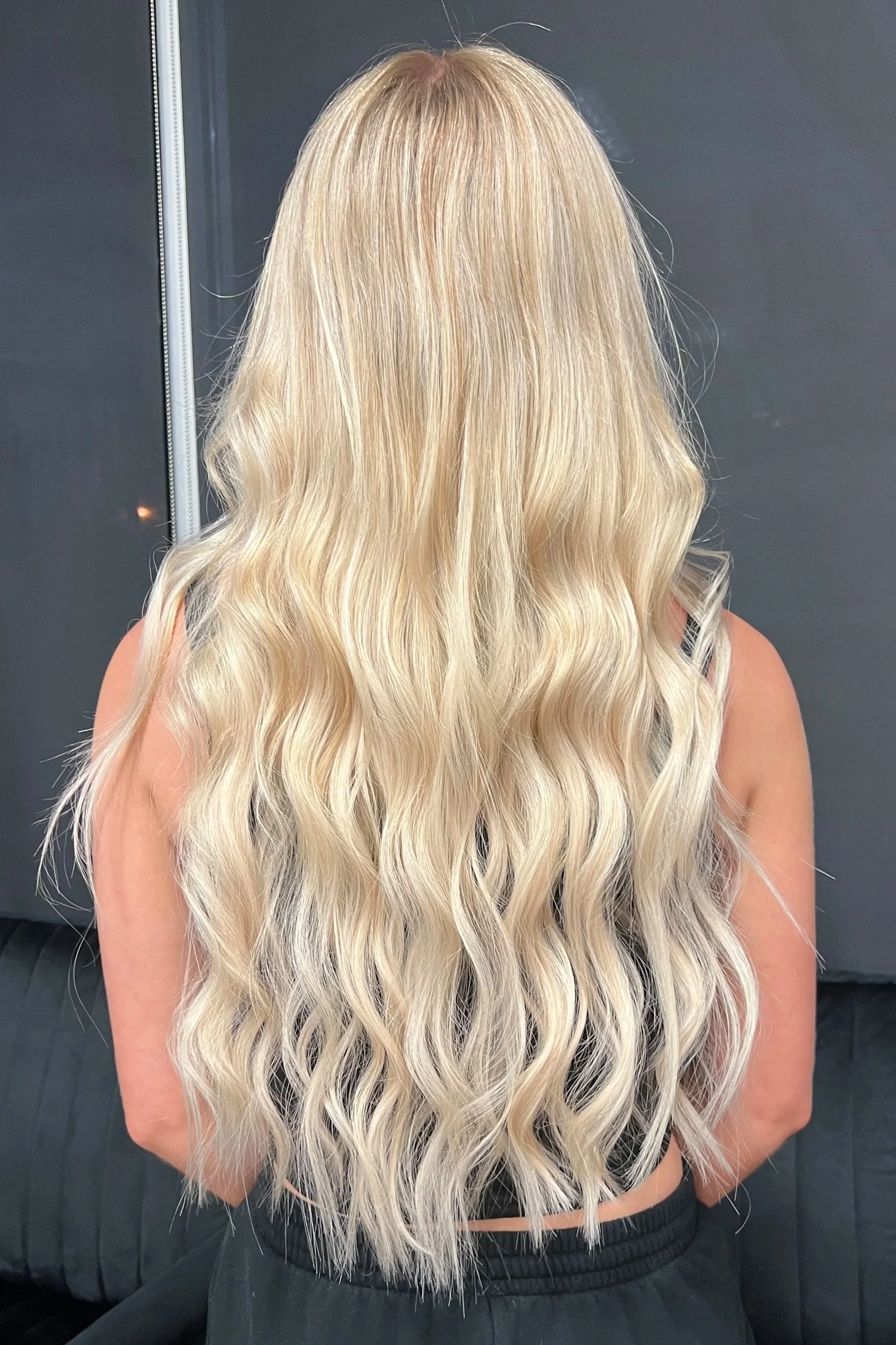 Back view of a woman with long, wavy blonde hair against a dark background.