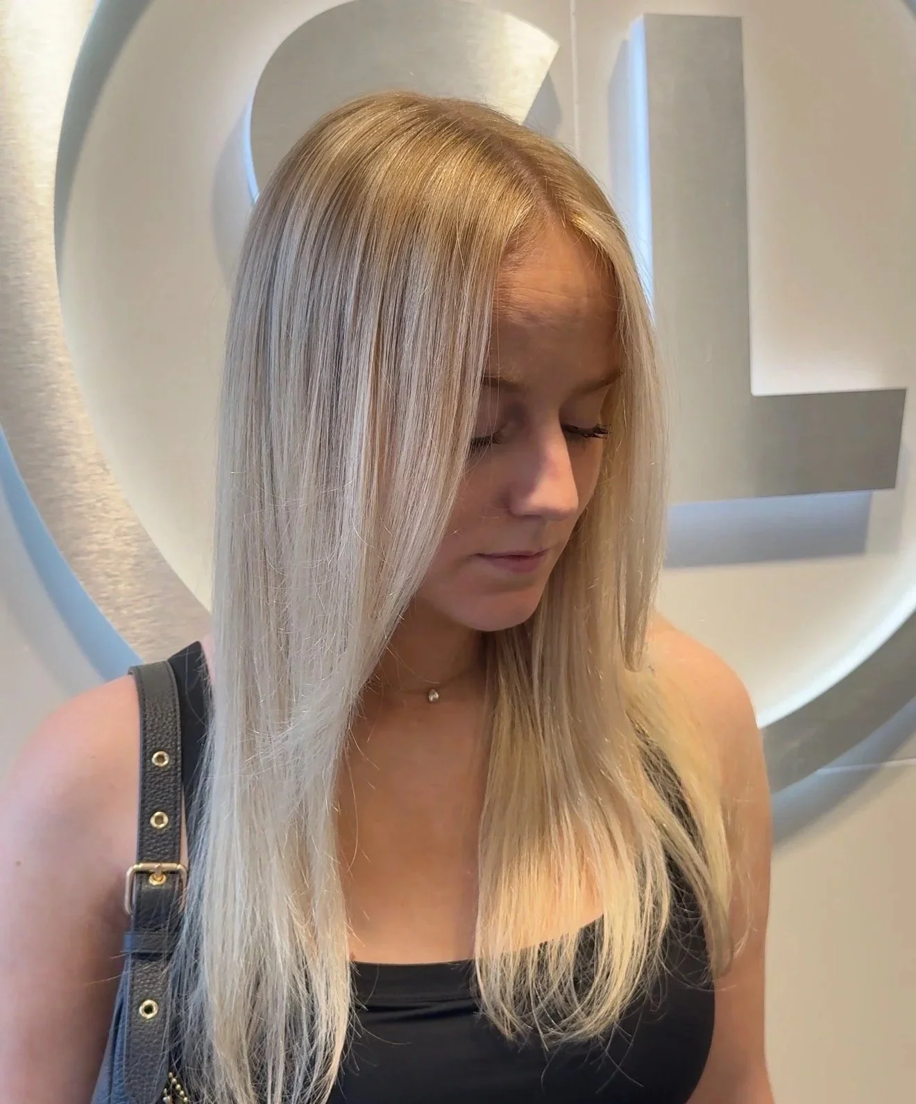 A woman with long, straight, blonde hair looking down, standing in front of a white background with a silver circular design.