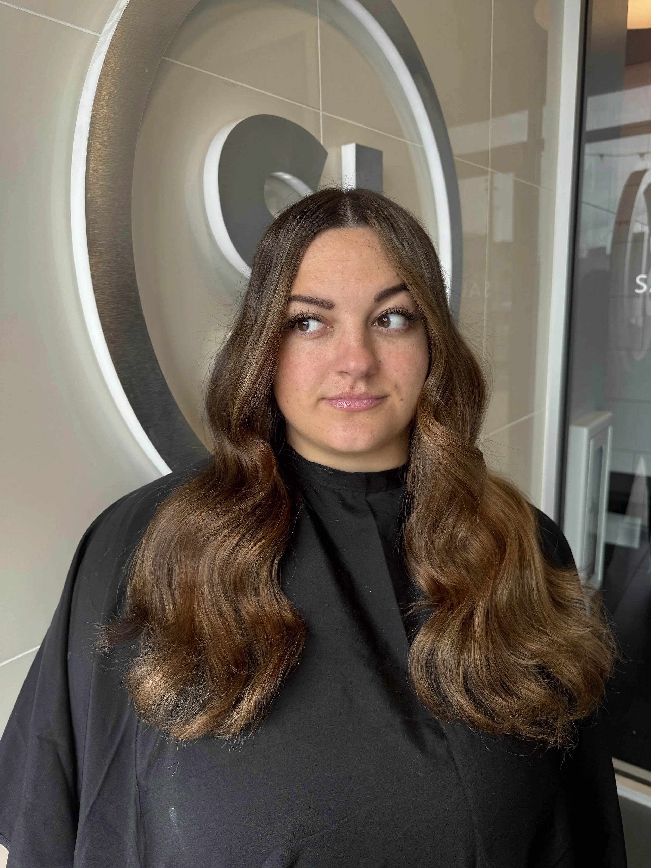 A woman with long wavy brown hair and light makeup looking slightly to the side in front of a large mirror in a salon or spa setting.