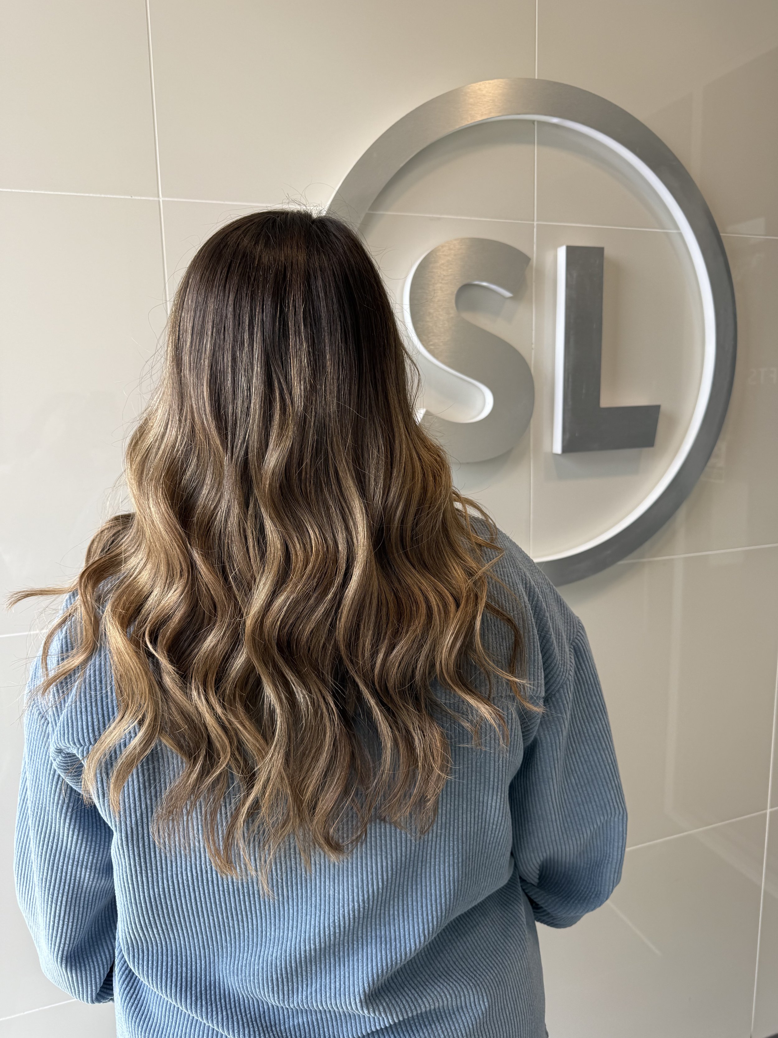 Back of a woman with long, wavy brown hair, wearing a blue ribbed sweater, standing in front of a wall with a large illuminated 'S' logo.