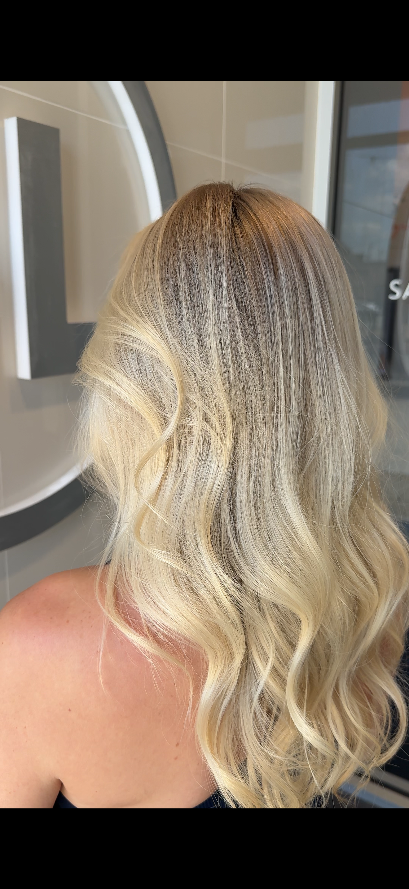 Back of a woman with shoulder-length, wavy blonde hair standing indoors near a mirror and wall.