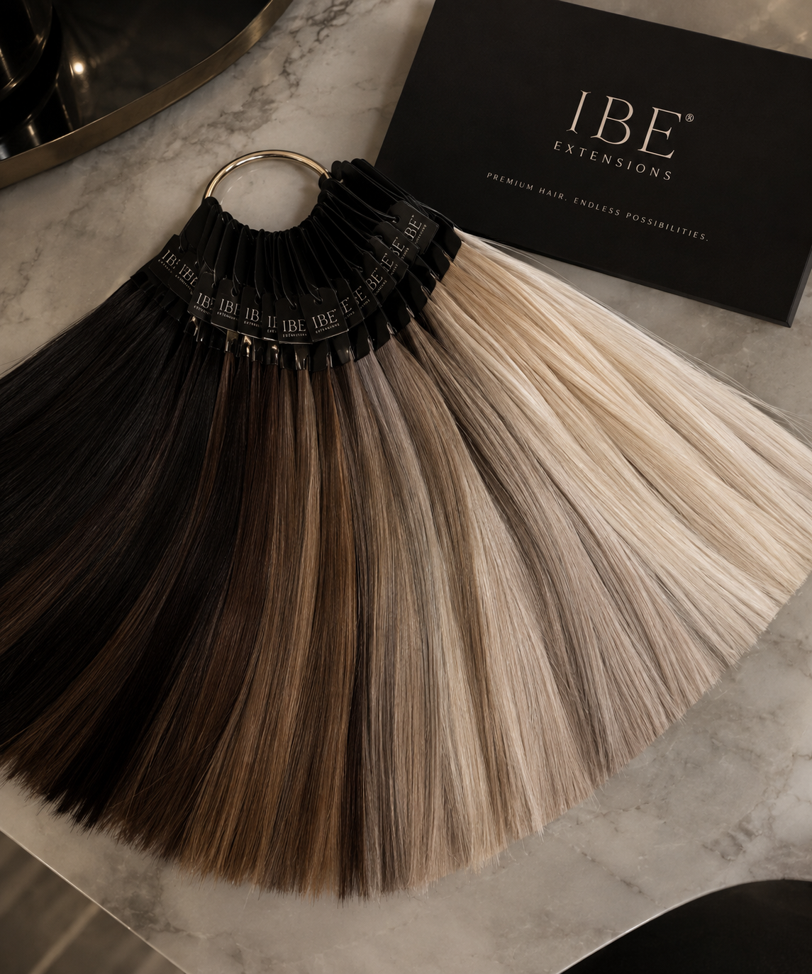 Hair color swatches in shades from dark brown to platinum blonde on a fan, with a black box labeled 'IBE Extensions' on a marble surface.