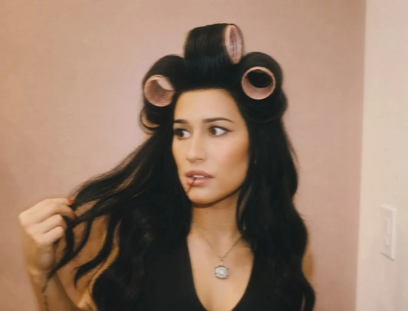 Woman with dark wavy hair using pink hair rollers, holding a strand of her hair, wearing a black top and a silver necklace, standing indoors against a plain beige wall.