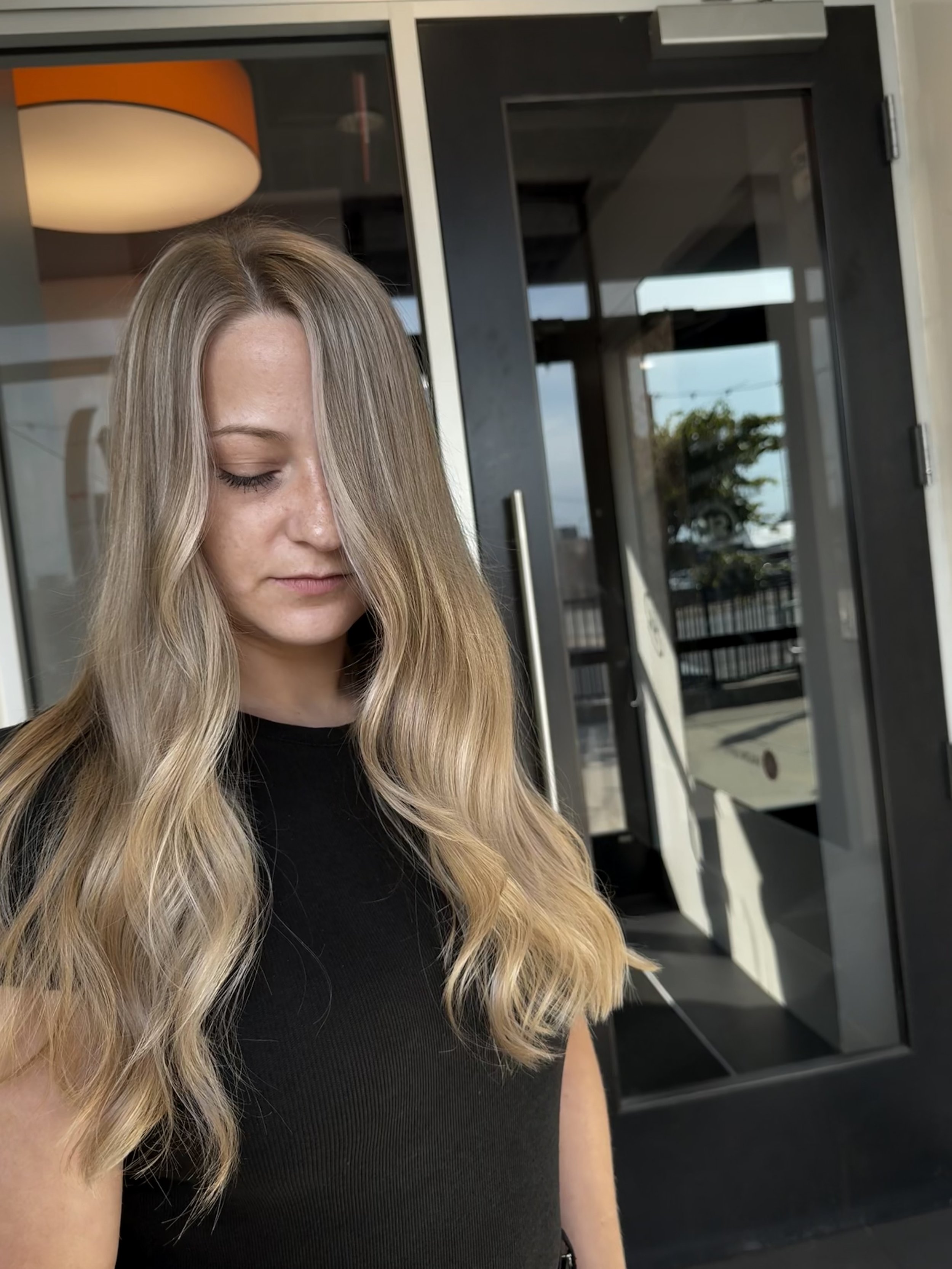 Young woman with long blonde hair and light skin standing outside near glass doors, wearing a black top.