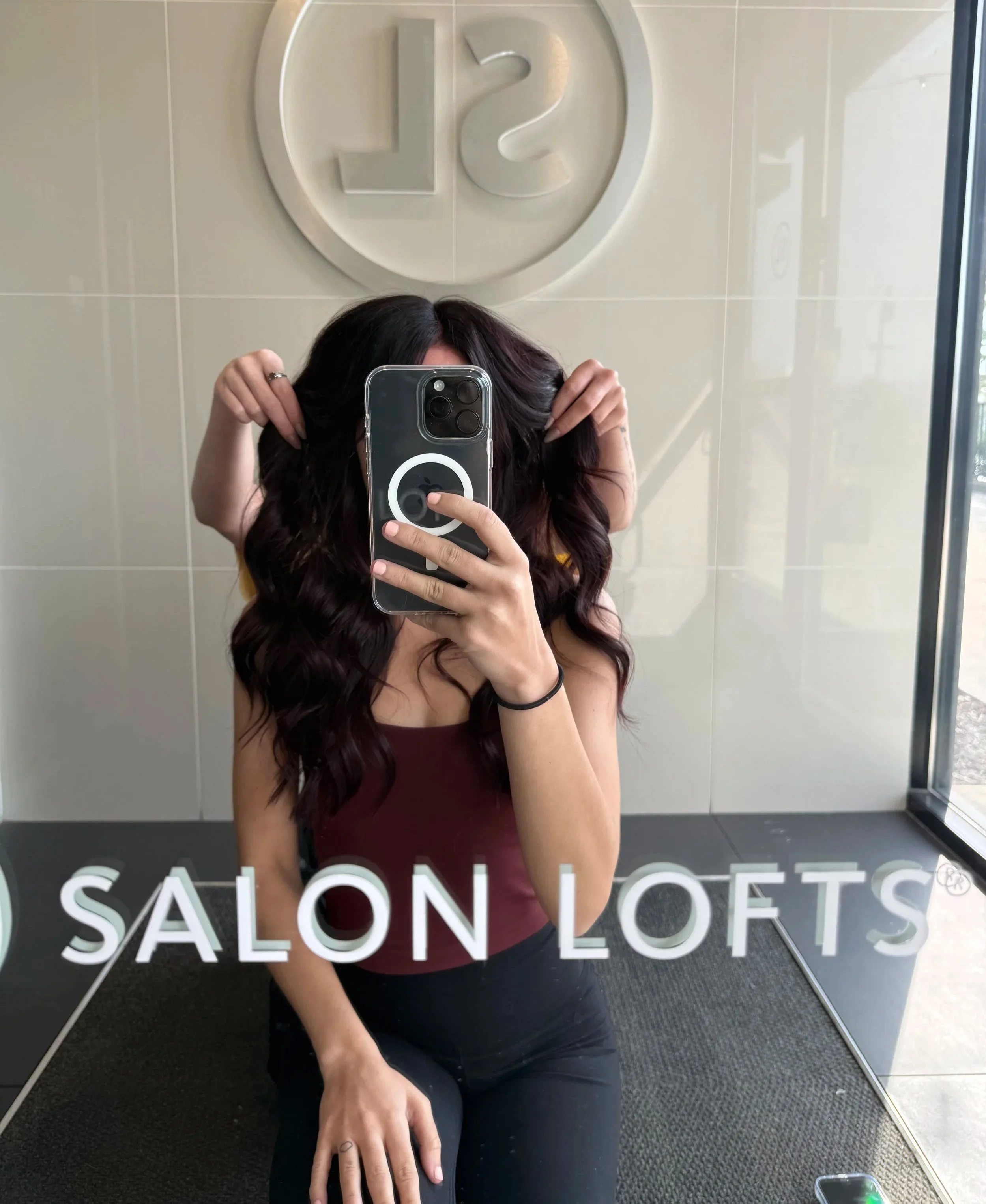 A person with dark wavy hair taking a mirror selfie. They are holding their hair with one hand and have a phone with a clear case and pop socket. The mirror has the words 'Salon Loc' written at the bottom, and a large circular logo with the numbers 12 is visible above. The background includes tiled walls and windows, indicating a salon or similar setting.