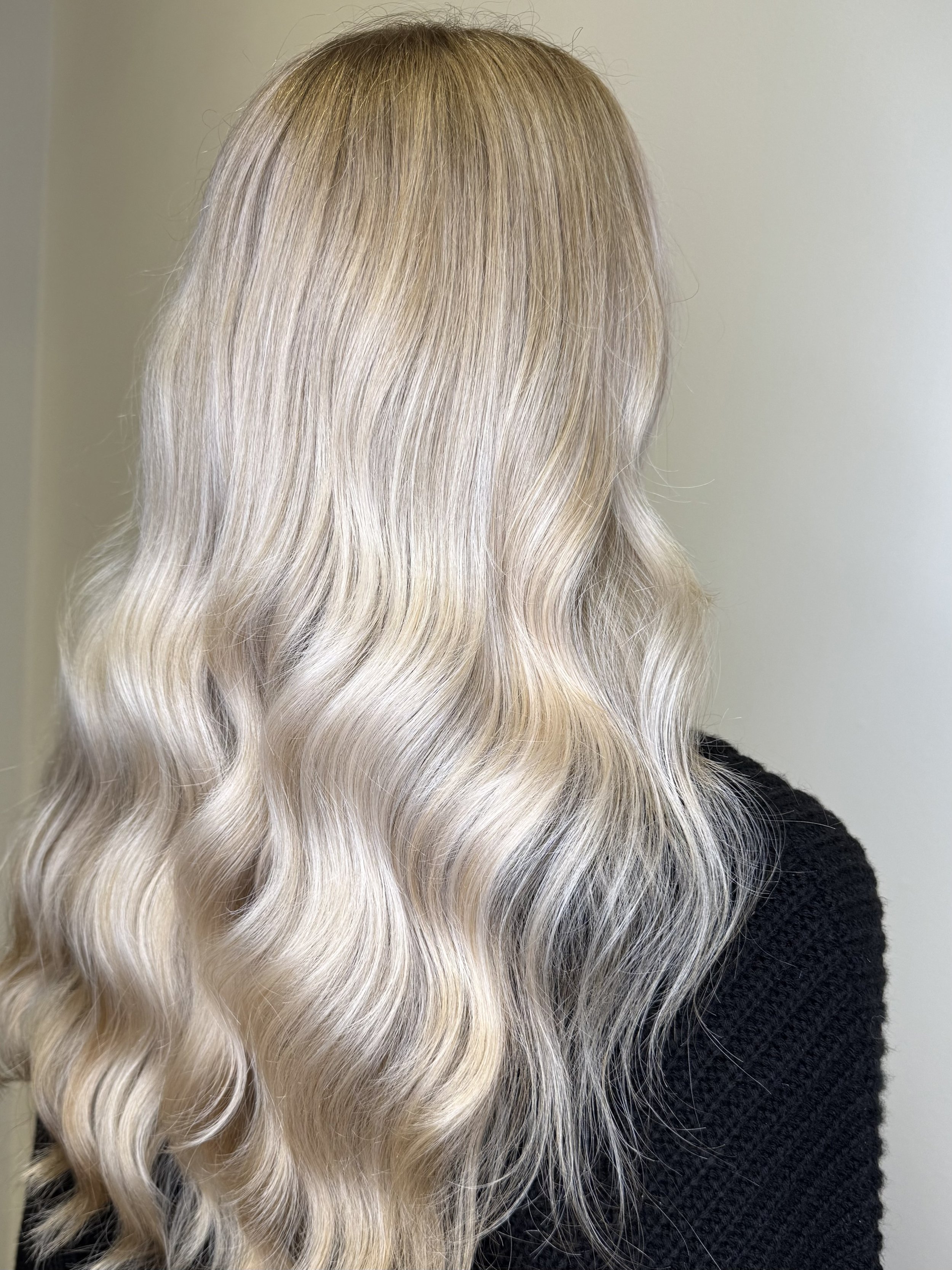 Close-up of long, wavy blonde hair with highlights, worn by a person wearing a black sweater, against a plain background.