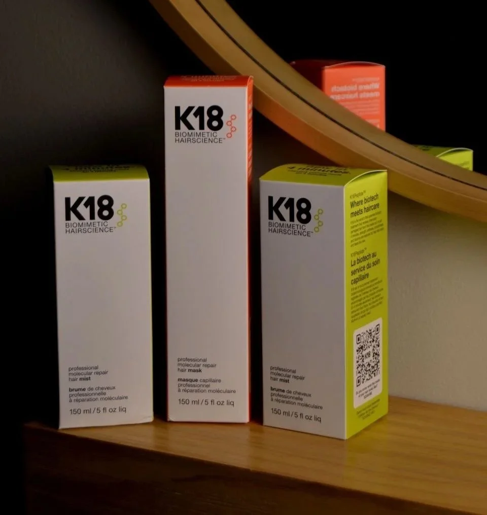 Three boxes of K18 Biometric Hair Science hair treatment products placed on a wooden surface near a mirror, with a curved mirror edge visible in the background.