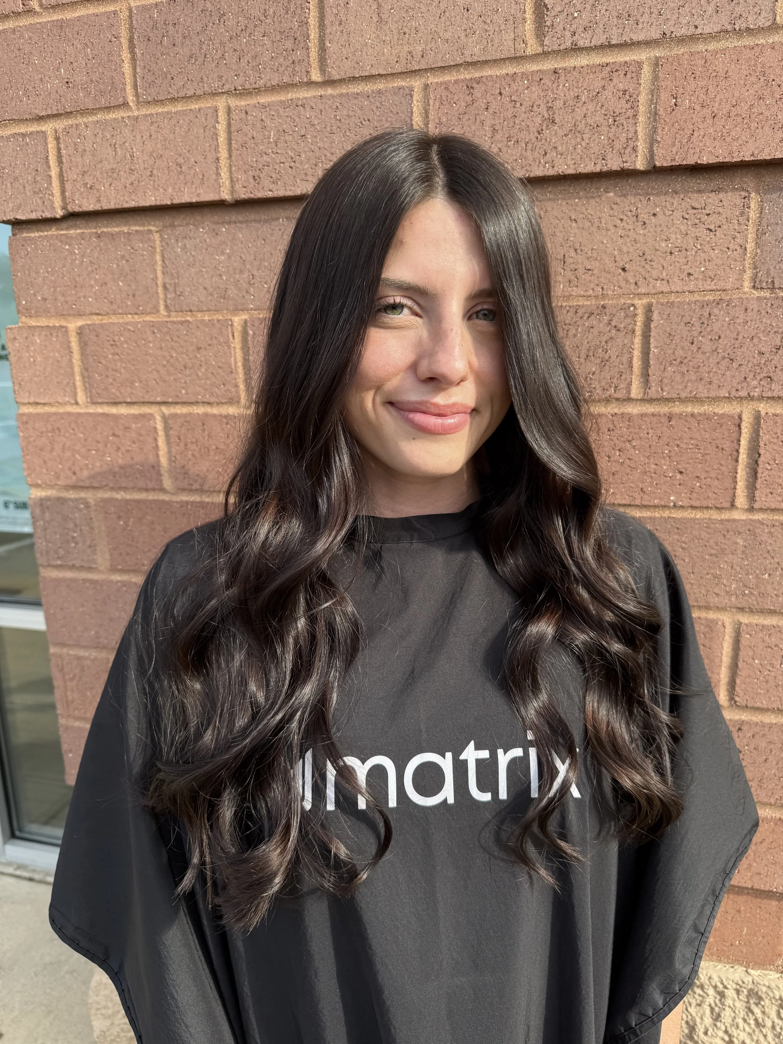A woman with long dark wavy hair and light green eyes standing in front of a brick wall.