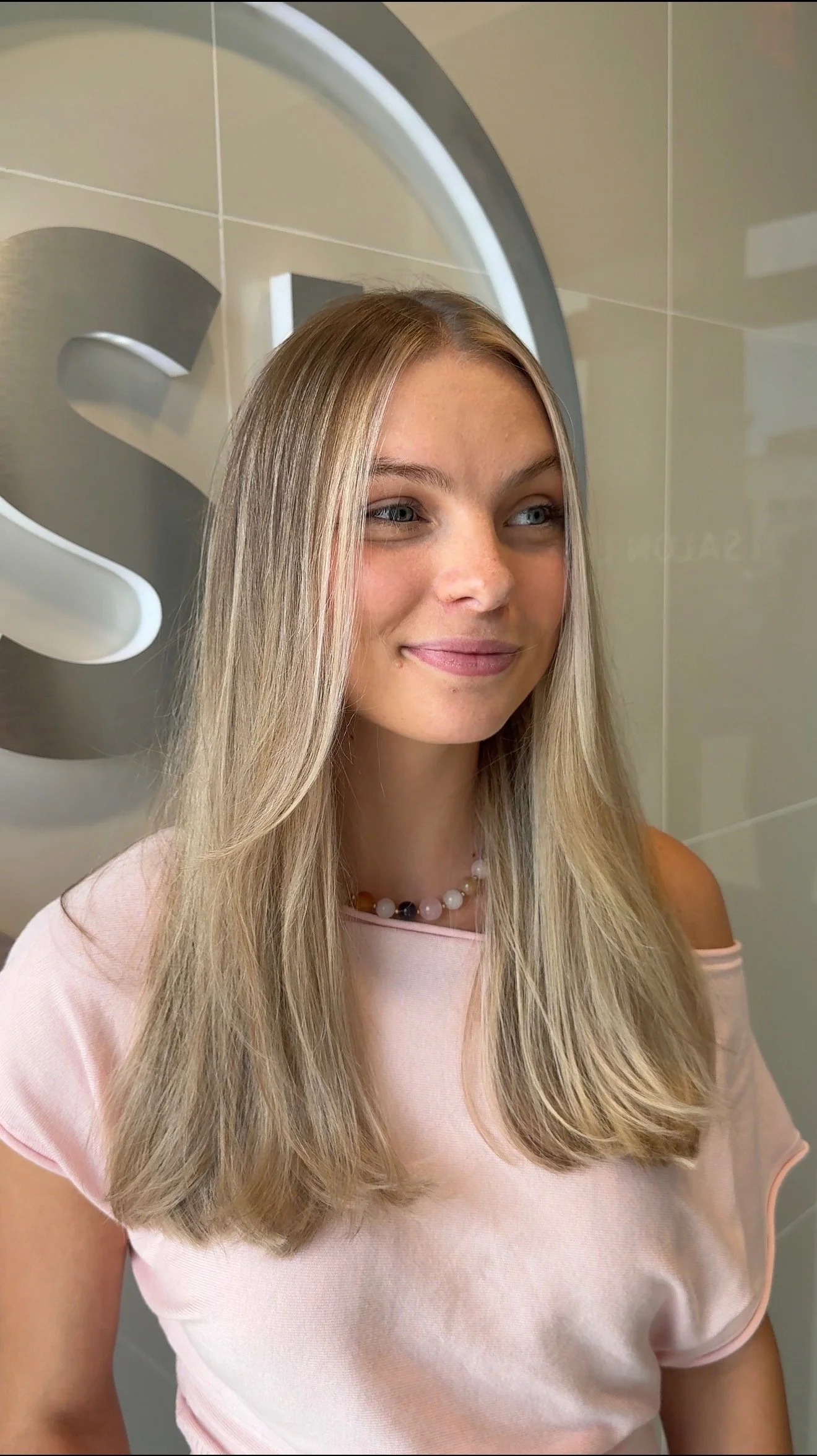 A smiling woman with long blonde hair, wearing a light pink top and a pearl necklace, posing indoors in front of a wall with a metallic logo or sign.